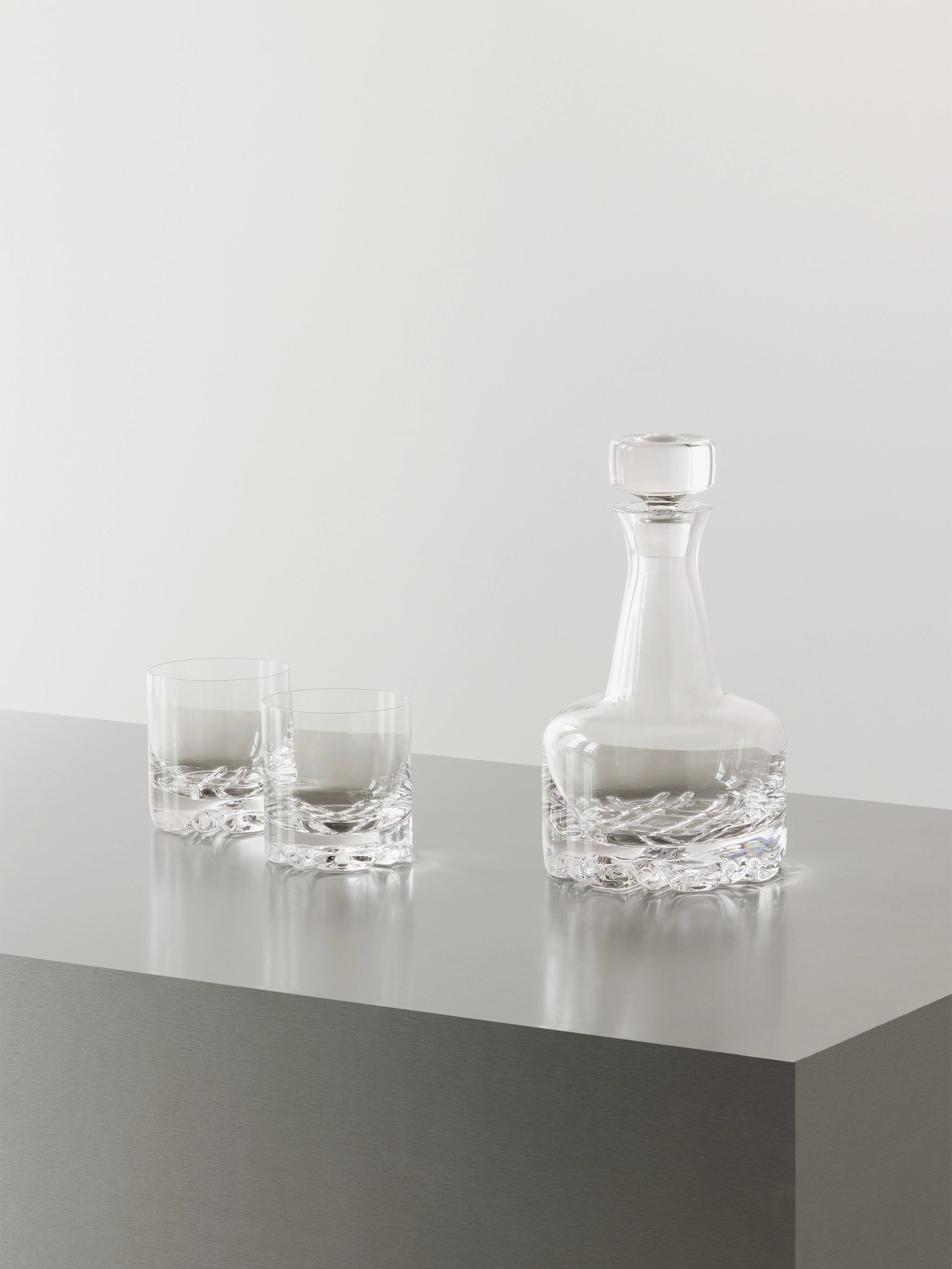 The Orrefors Erik 3-Piece Set includes a 26oz decanter and two 12oz double old fashioned glasses, all showcasing sleek Scandinavian design, arranged on a gray metallic surface with a plain light background.