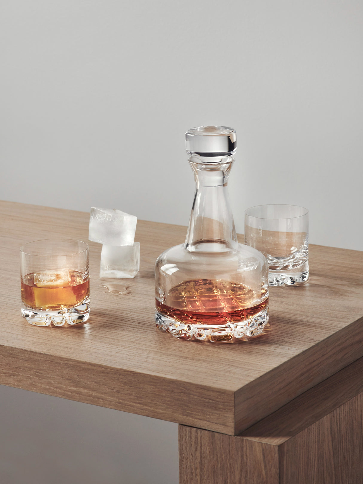 The Orrefors Erik 3-Piece Set includes a 26oz glass decanter filled with whiskey, placed on wood next to two 12oz double old fashioned glasses and three ice cubes, reflecting timeless Scandinavian design.