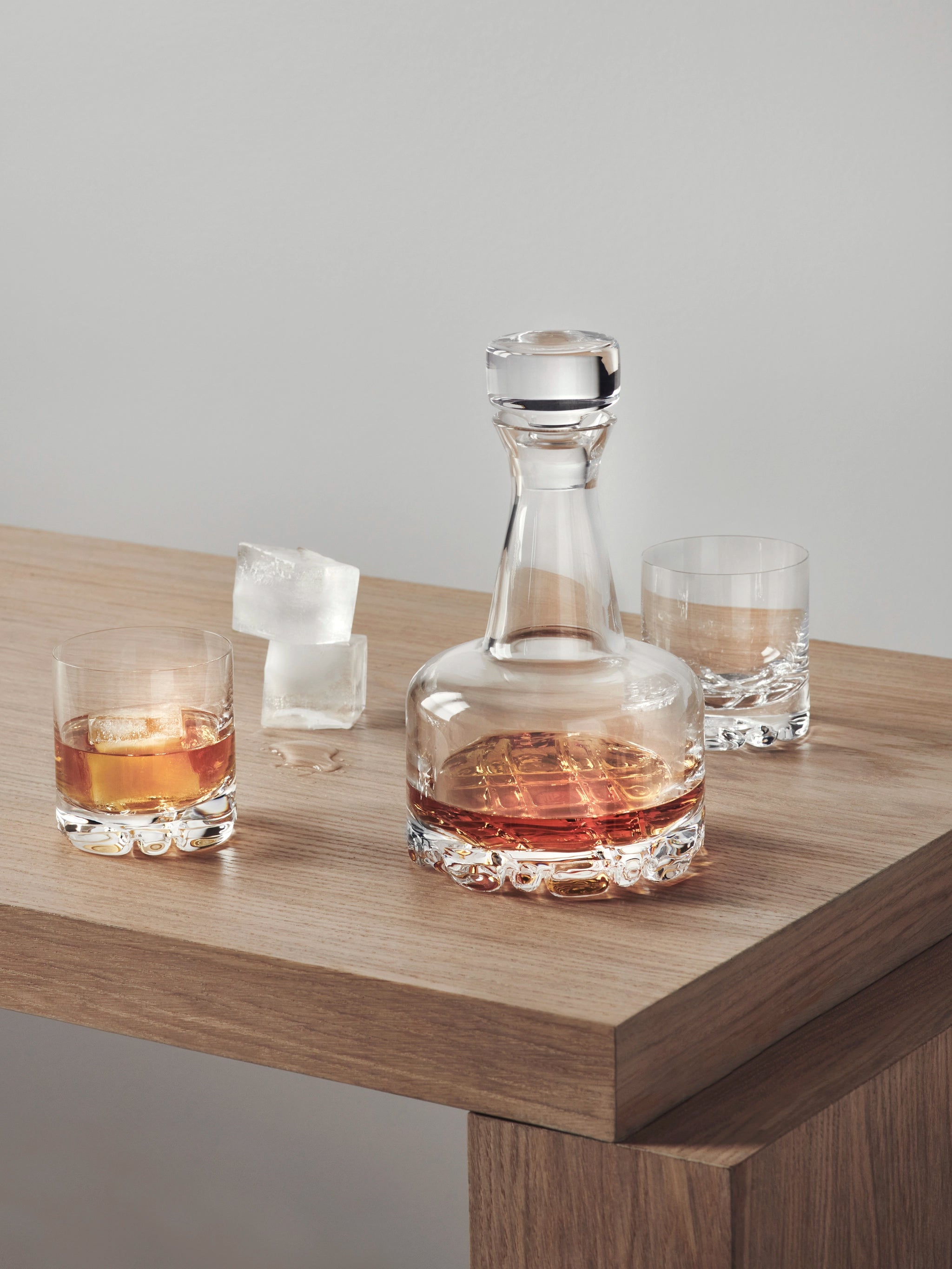 The Orrefors Erik 3-Piece Set includes a 26oz glass decanter filled with whiskey, placed on wood next to two 12oz double old fashioned glasses and three ice cubes, reflecting timeless Scandinavian design.