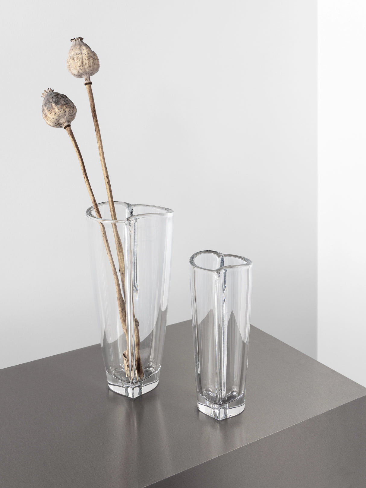 Two Orrefors Heart Bud Vases sit on a gray surface, with the larger one holding two dried poppy seed pods.