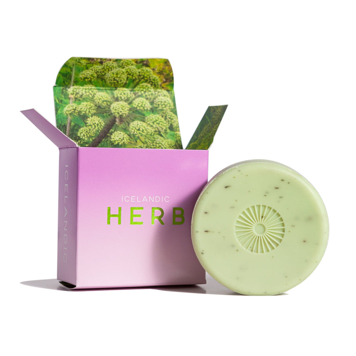 A round green soap featuring speckles and infused with chlorophyll and Angelica Herb antioxidants is partially outside its open pink and green Hallo Iceland Angelica Herb Bar Soap box, adorned with a floral pattern.