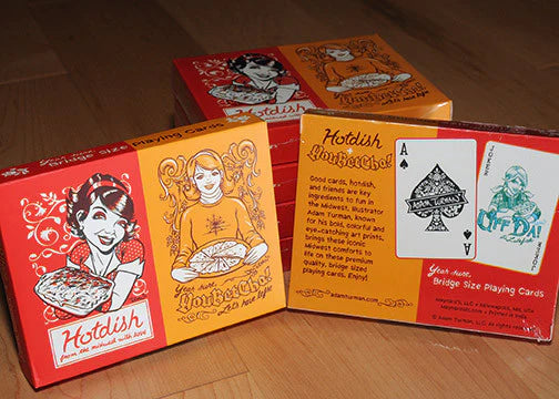 Two boxes of Playing Cards: Hotdish/YouBetCha Bridge Size Playing Cards are shown, with one box open to display a bridge-size deck featuring an Ace of Spades and a green Joker card designed by Adam Turman.