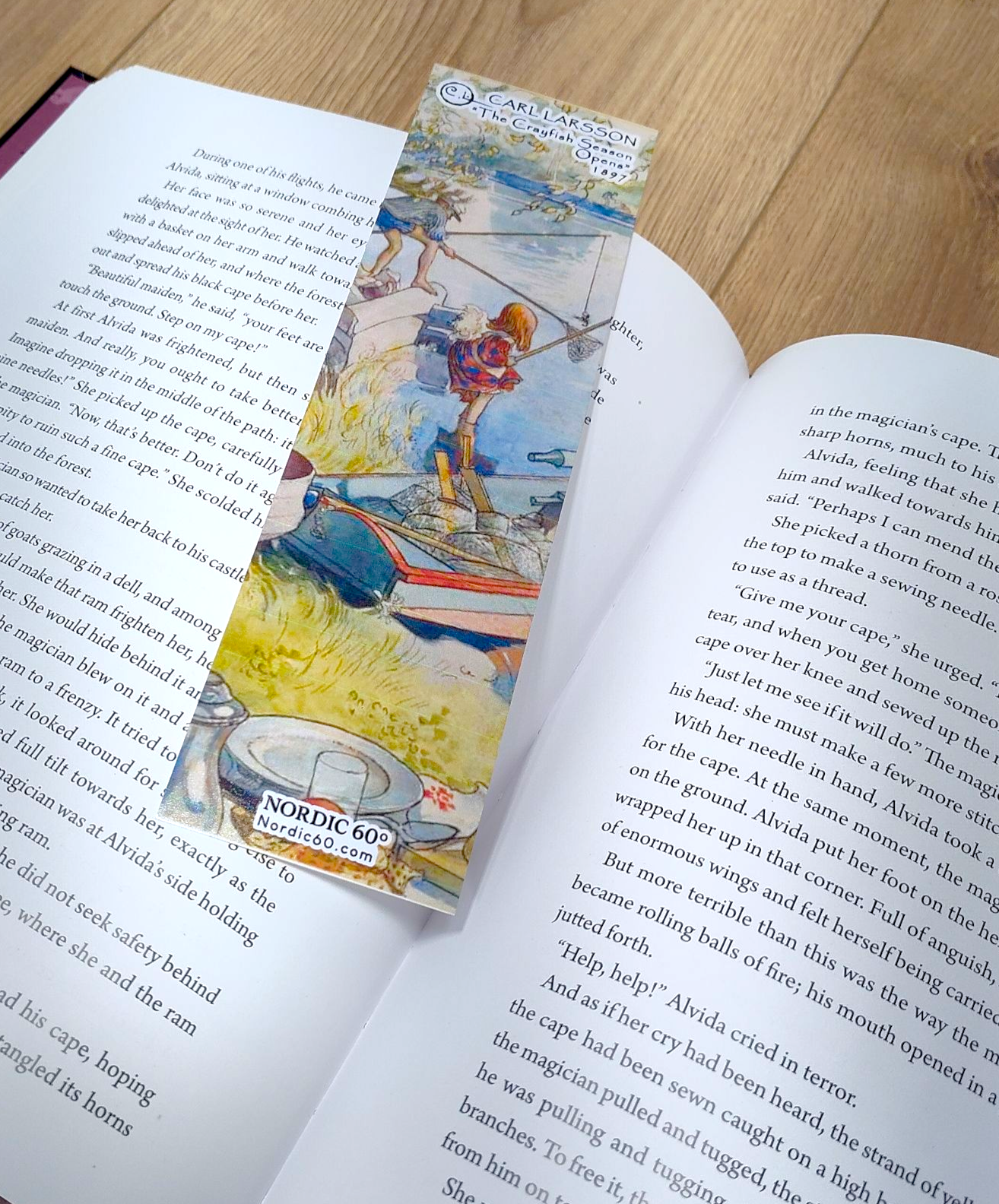 A Carl Larsson “The Crayfish Season Opens” bookmark (8.5" × 2.5") with a vivid fishing scene illustration sits on an open book atop a wooden surface, capturing the charm of Swedish summer art.