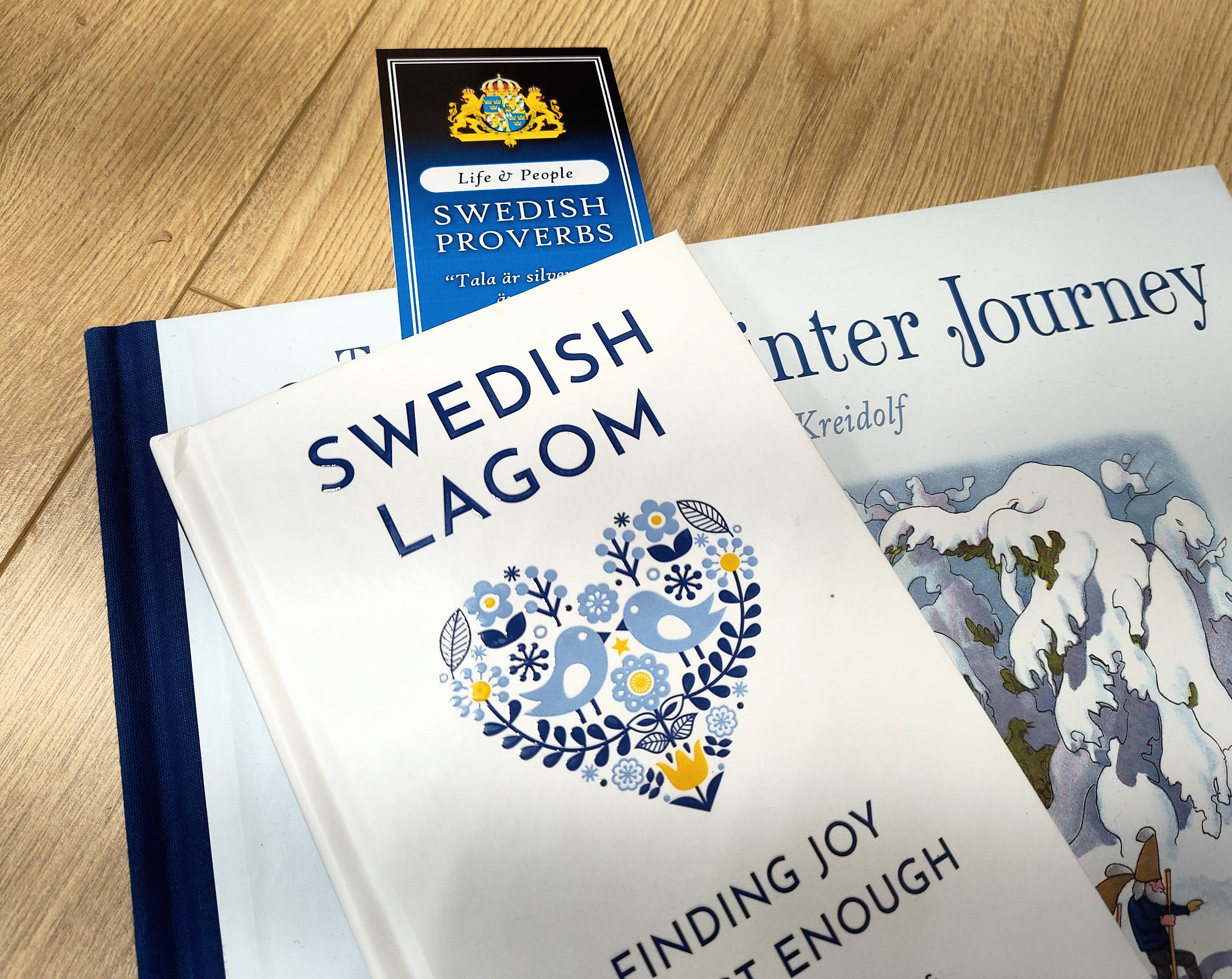 Three items on a wooden surface: the book “Swedish Lagom,” the bookmark “Swedish Proverbs ‘Life & People’” (2.5" × 8.5”), and “The Winter Journey” with an illustrated snowy scene on its cover.