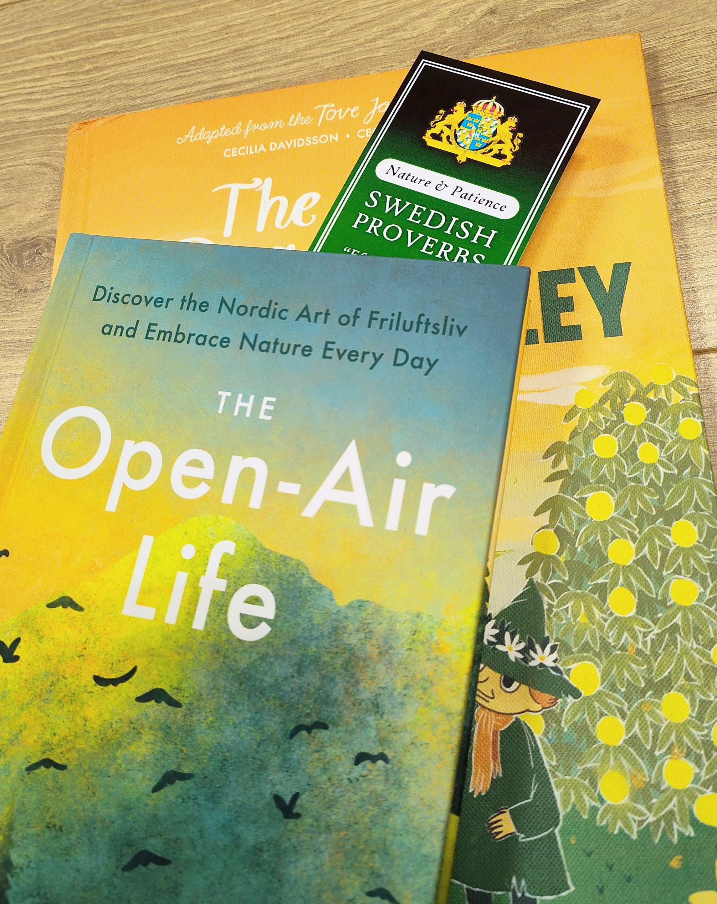Three books are stacked on a wooden surface. The top book is titled The Open-Air Life, and a Bookmark: Set of 4 Swedish Proverbs (2.5" × 8.5") marks a page in the second book below, highlighting Scandinavian wisdom.