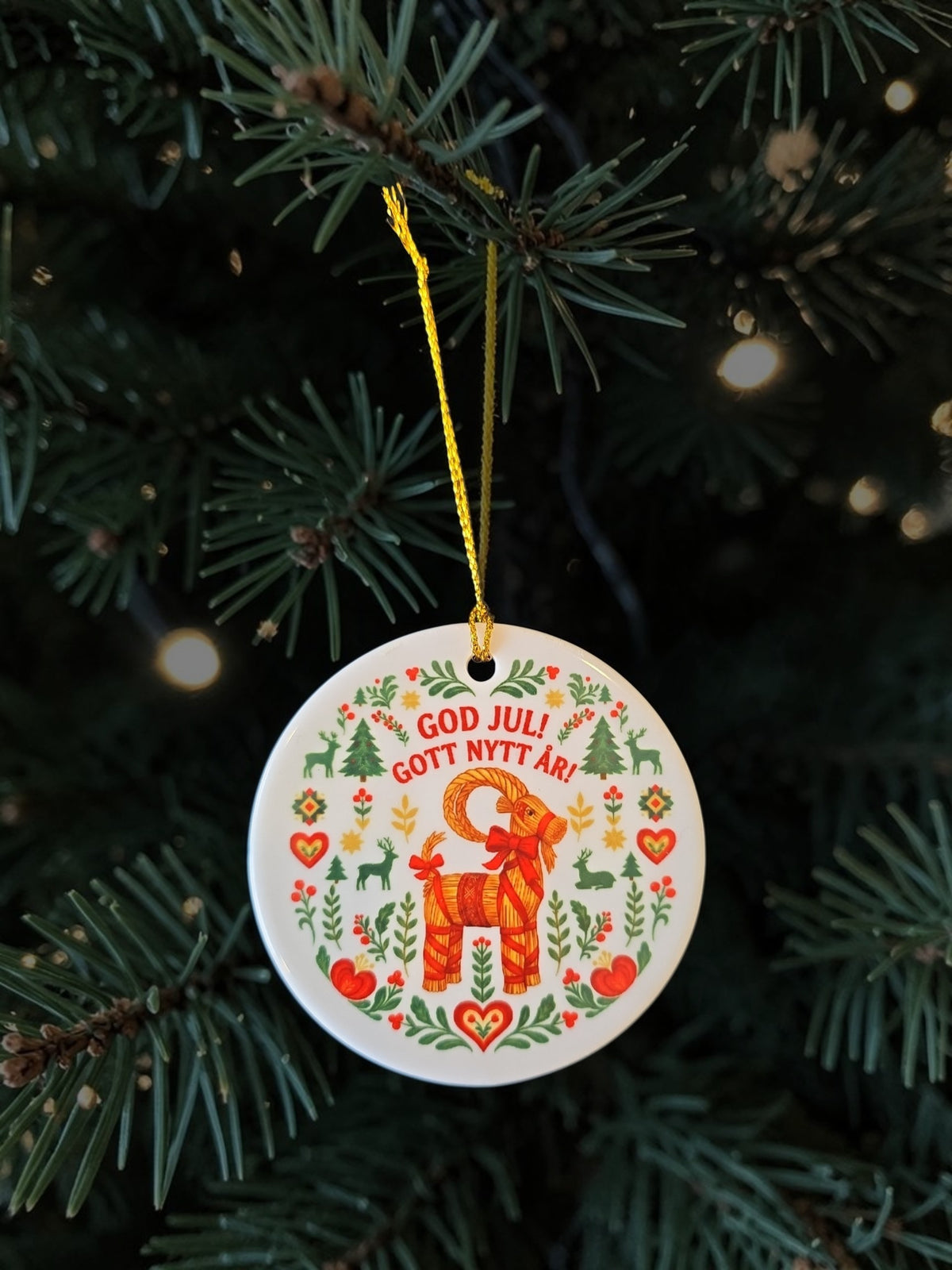 A round white ceramic Christmas ornament featuring a red ram illustration and Swedish text 'GOD JUL! GOTT NYTT ÅR!' surrounded by green leaves and red hearts, hanging from a yellow string on a pine tree branch.