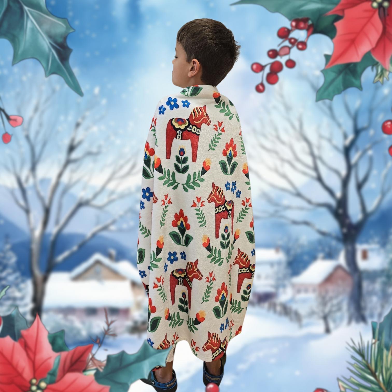 A young boy wrapped in a Dala Horse Children's Blanket 29" x 40" stands facing away before a snowy winter village, with holly and berry decorations framing the scene.