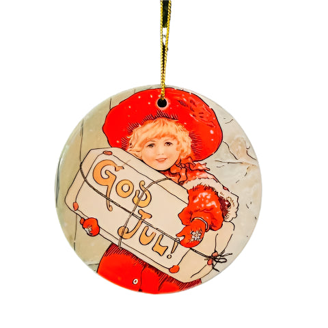The Ornament: GOD JUL! Girl Holding Sign is a round porcelain piece featuring an illustration of a child in red holding a sign that says "God Jul." It hangs beautifully on a green tree, infusing unique charm into your holiday decoration project.