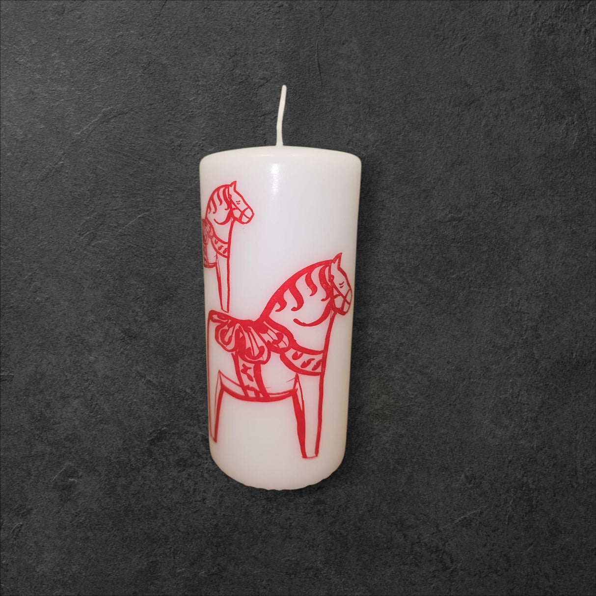 The Red Dala Horse Pillar Candle by Logum Kloster Lys in Denmark features red, stylized Swedish Dala Horses on a white candle, creating a striking contrast.