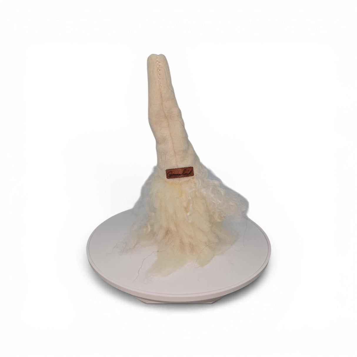 The Gnome: "Foot Tomte" by Talla Imports, featuring a white sculpted hat, woolly base, and clay nose and feet, stands upright on a round white platform against a plain background. This 6-inch handcrafted gnome from Gumselids of Sweden also features a distinctive brown band accent.