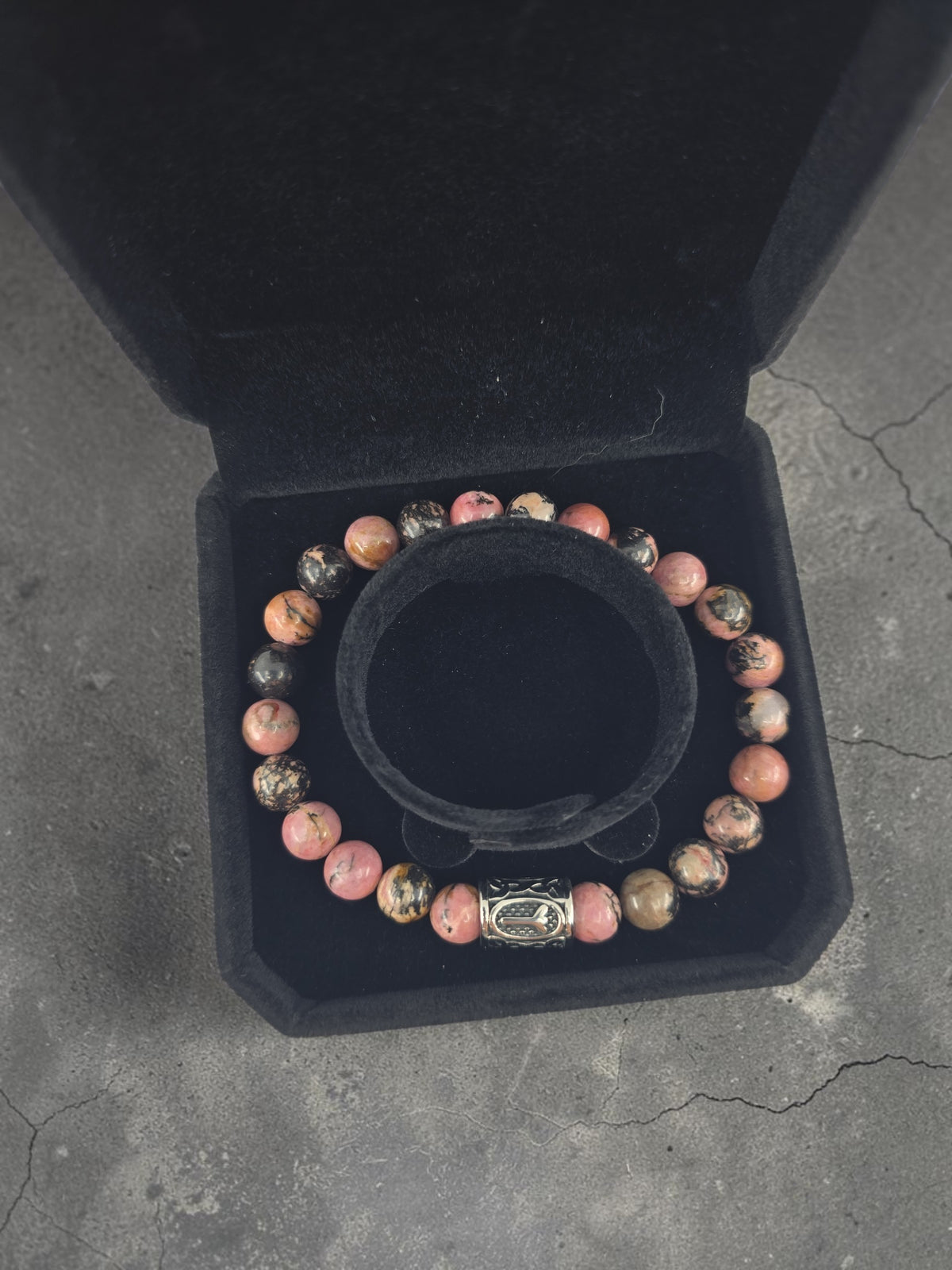 Pink and black bracelet featuring an Algiz rune and silver clasp, displayed in an open black velvet jewelry box on a gray cracked surface.