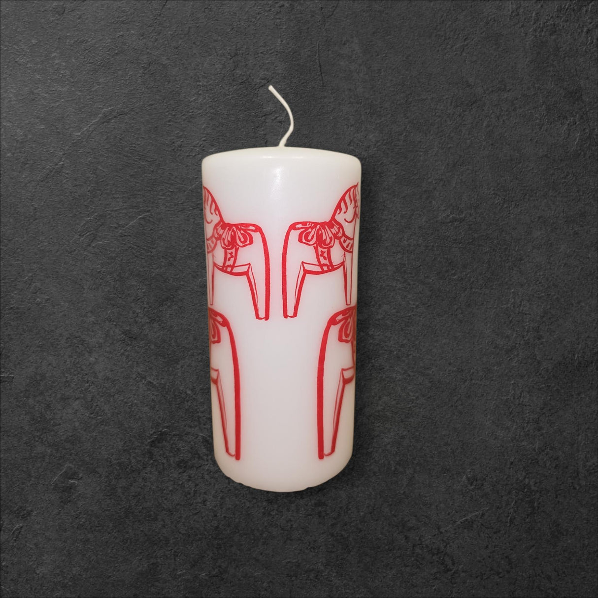 A Logum Kloster Lys Red Dala Horse Pillar Candle from Denmark is shown upright, featuring red line art of dala horses on its white surface against a dark textured background.