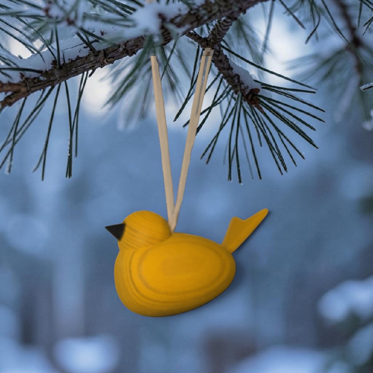 The Swallings Wooden Yellow Swedish Bird Hanging Ornament, made in Sweden, hangs from a pine branch by two strings against a snowy, blurred outdoor background.