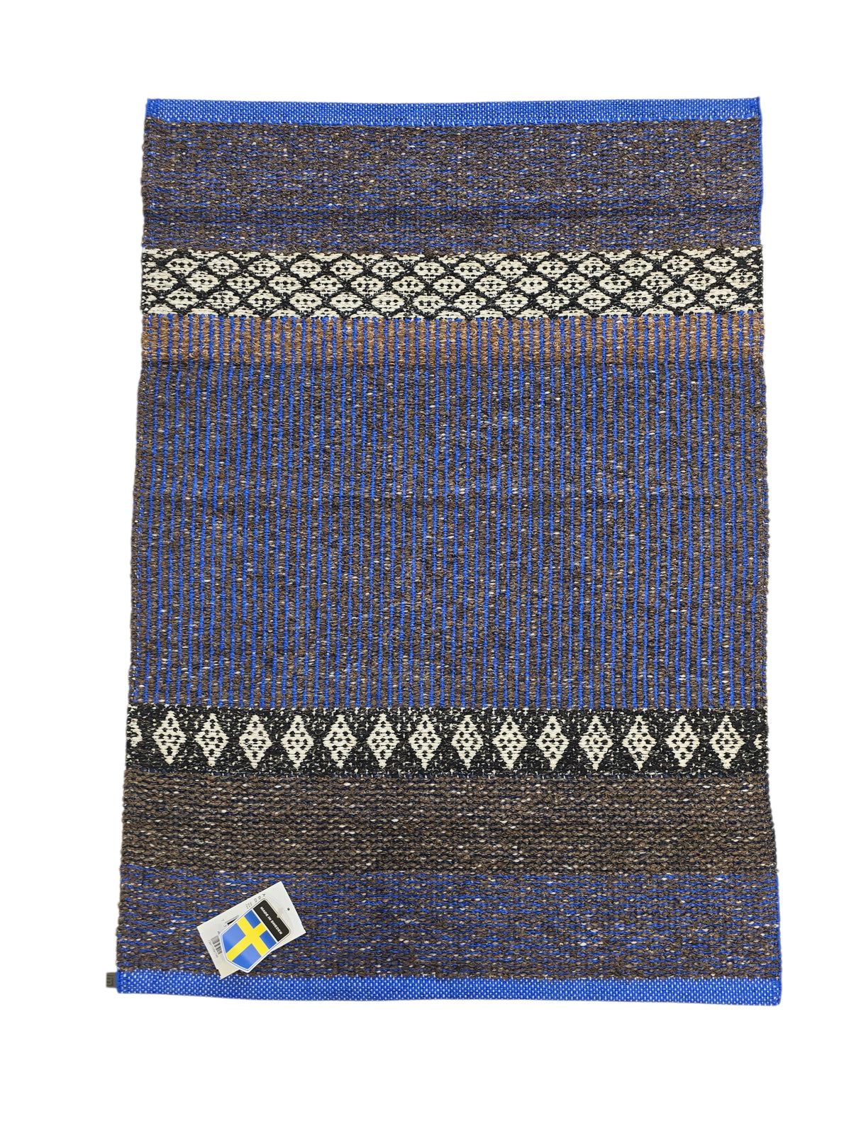 The Horredsmattan Mixed Forest Cobalt rug is a rectangular Swedish cotton/vinyl rug with blue, brown, and black patterns, white geometric designs, and a small Swedish flag tag in the bottom left corner.