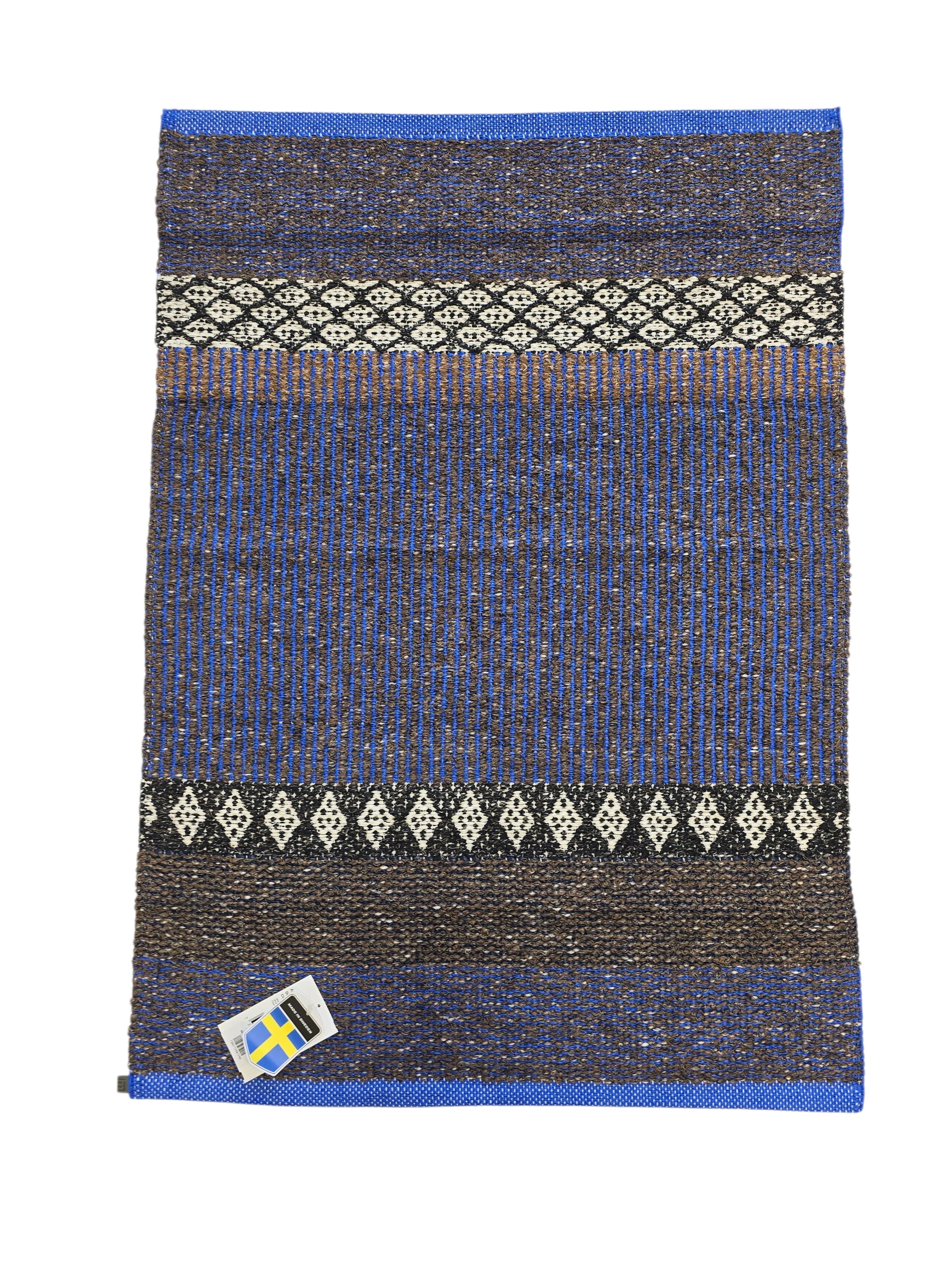The Horredsmattan Mixed Forest Cobalt rug is a rectangular Swedish cotton/vinyl rug with blue, brown, and black patterns, white geometric designs, and a small Swedish flag tag in the bottom left corner.