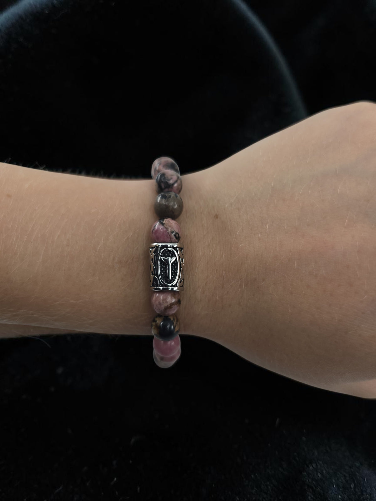 A hand wearing a beaded bracelet with pink and brown stones and a silver Algiz rune charm, shown against a black background.