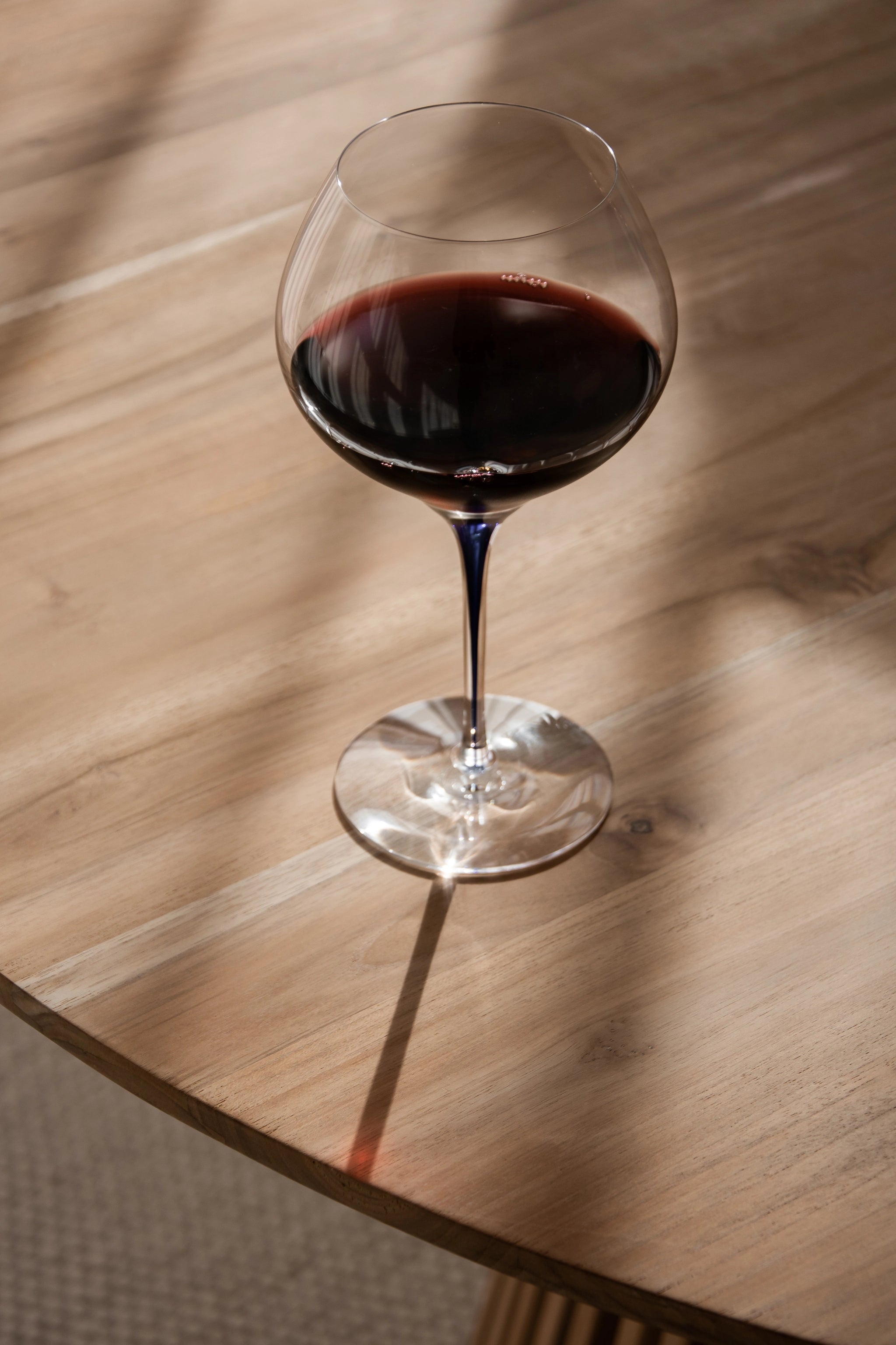 A glass of red wine catches the sunlight on a wooden table, its elegance highlighted by the Orrefors Intermezzo Blue Bouquet 20oz glass with a clear bowl and blue stem, designed by Erika Lagerbielke.