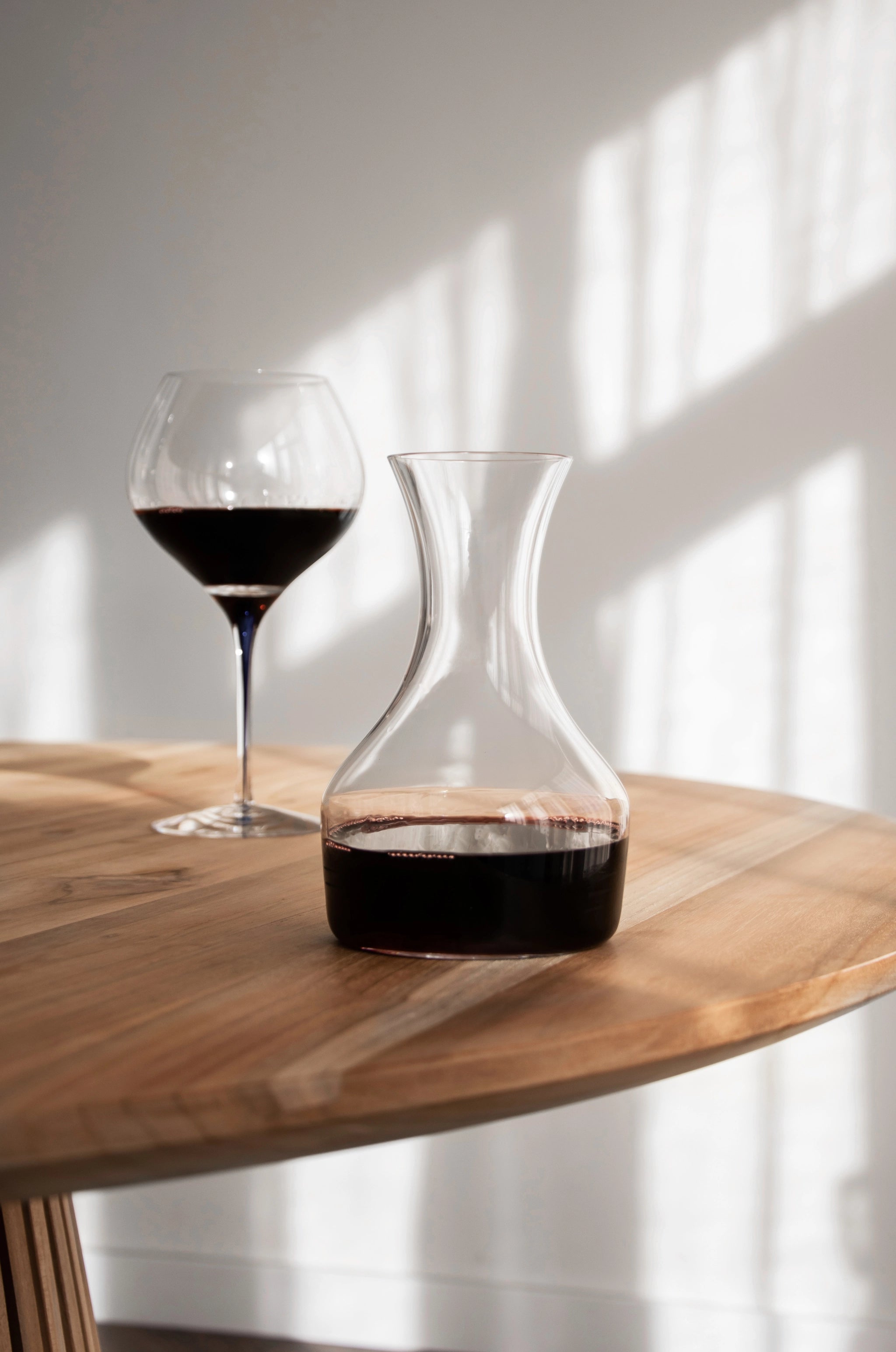 An Orrefors Intermezzo Blue Bouquet 20oz glass with a blue stem by Erika Lagerbielke, filled with red wine, sits on a round wooden table in natural sunlight.
