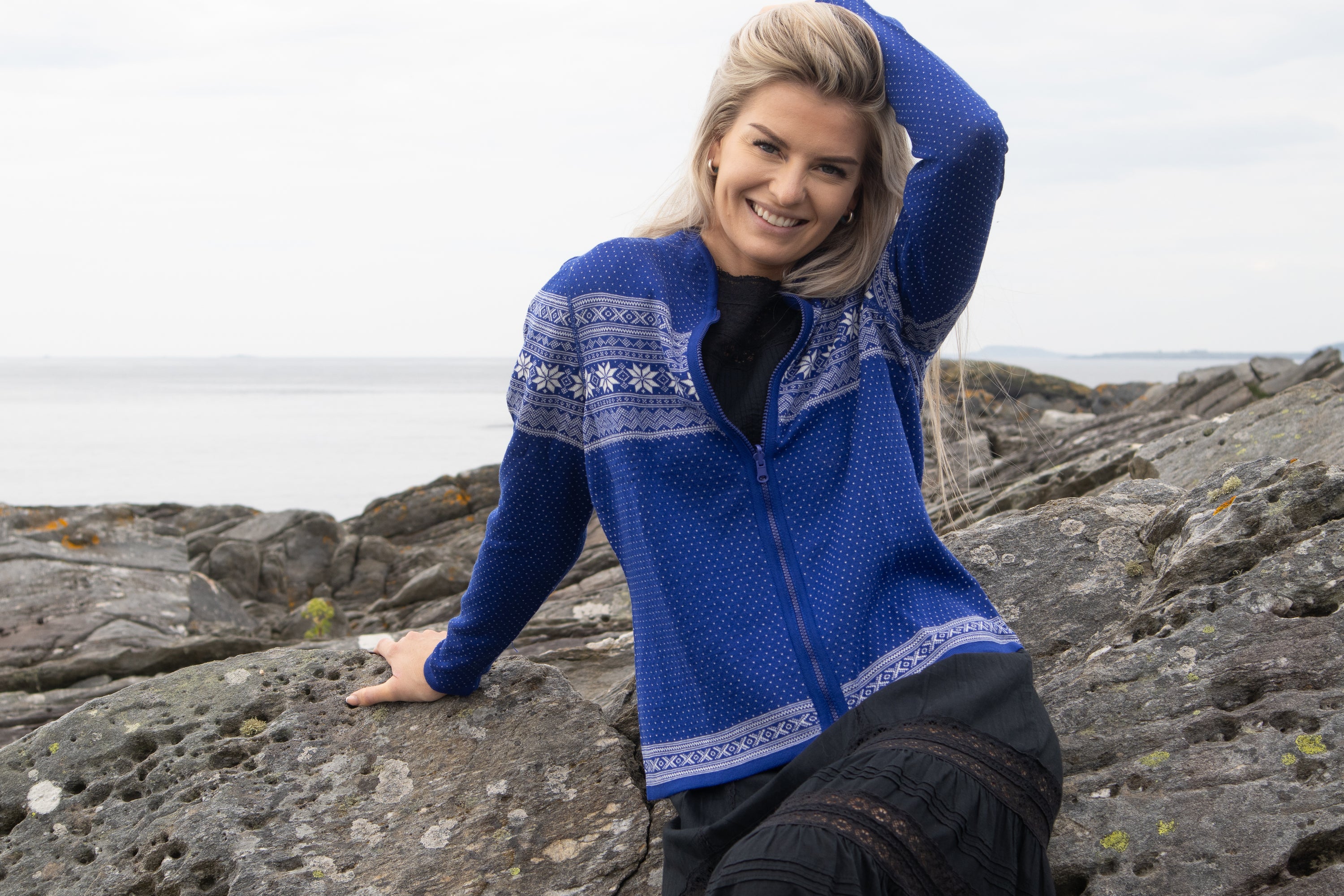 A woman in the Northern Story Setesdal Royal Blue Norwegian Design Wool Cardigan sits on rocky seaside terrain, smiling and resting her left hand on her head.