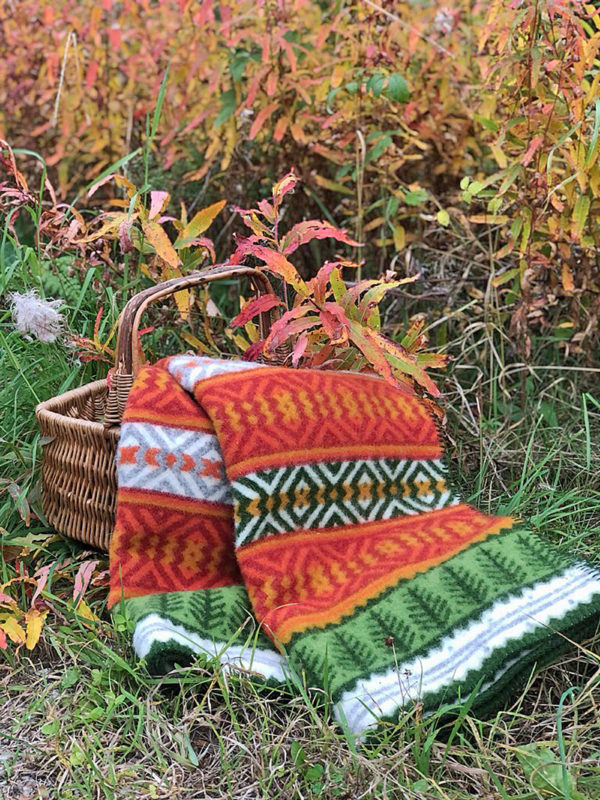 A woven basket rests on grass next to autumn plants, draped with the Mora Blanket—100% Norwegian lamb’s wool by Kerstin Landström Design—in green, orange, white, and red.
