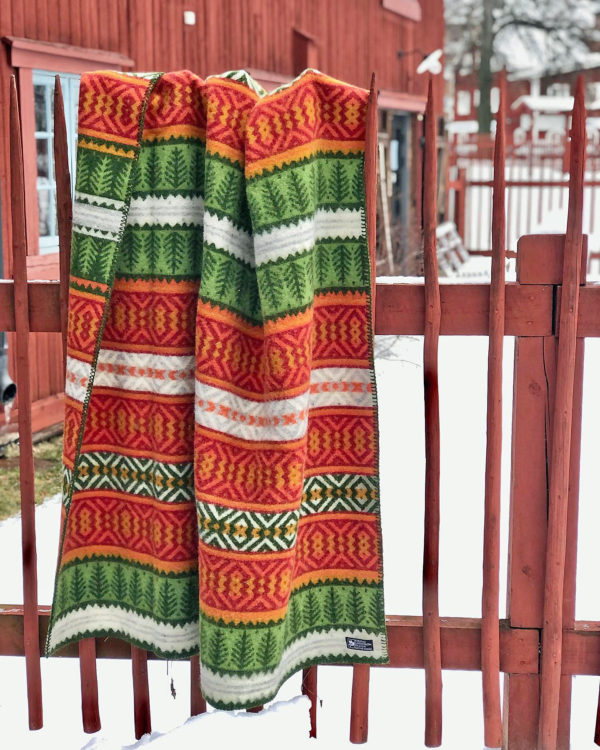 The Mora Blanket—100% Norwegian lamb’s wool by Kerstin Landström Design—featuring red, green, yellow, and white Scandinavian patterns, hangs over a wooden fence with snow and a red building in the background.