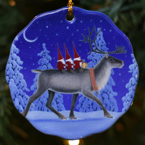 The Ornament: Tomte Riding Reindeer on Round Porcelain Ornament features artwork by Eva Melhuish, showing three gnomes riding a reindeer through a snowy forest at night with snow-covered trees and a crescent moon.