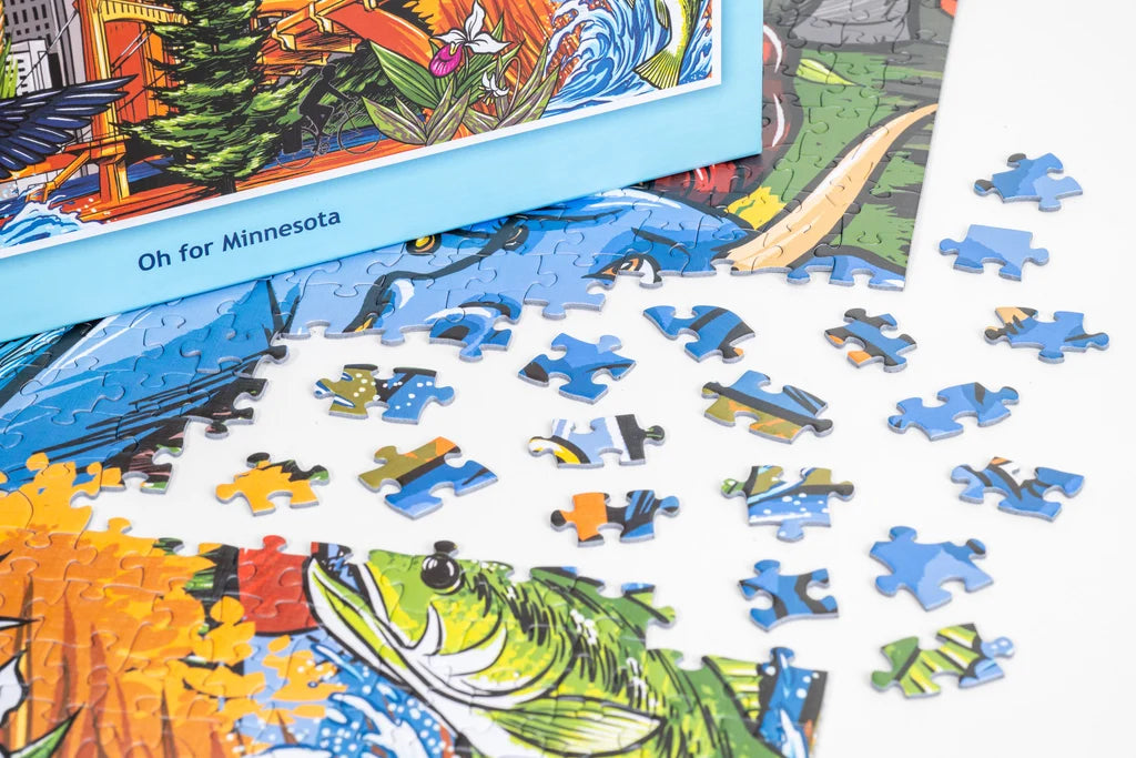 Partially assembled puzzle featuring colorful Minnesota-themed artwork, with loose pieces and the box lid visible on a white surface.