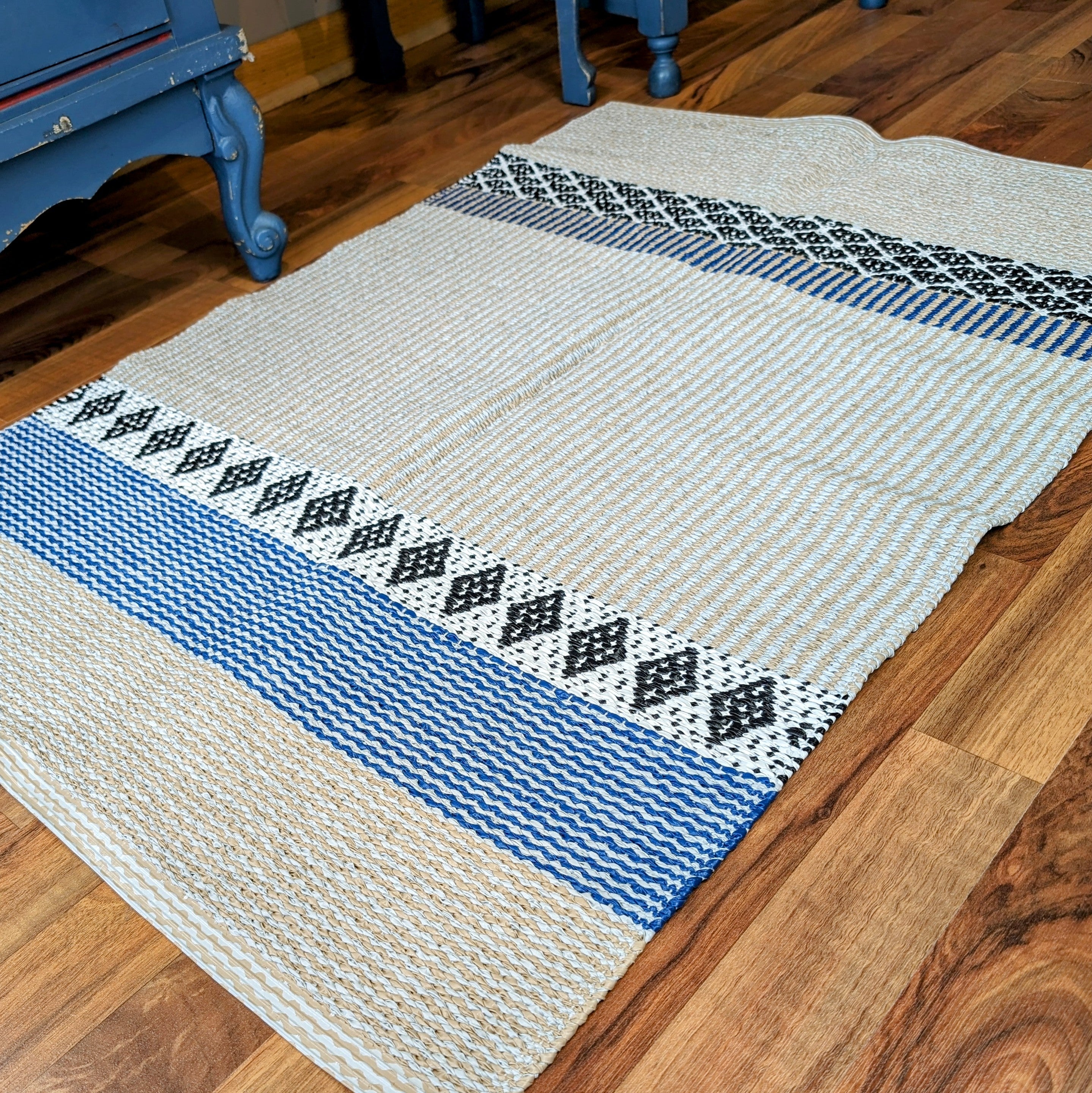 The Savanne Blue Vinyl Rug is gracefully displayed on a wooden floor near blue furniture, featuring intricate blue, black, and white patterns. This Horredsmattan creation provides a stylish touch and is maintenance-free for effortless living.