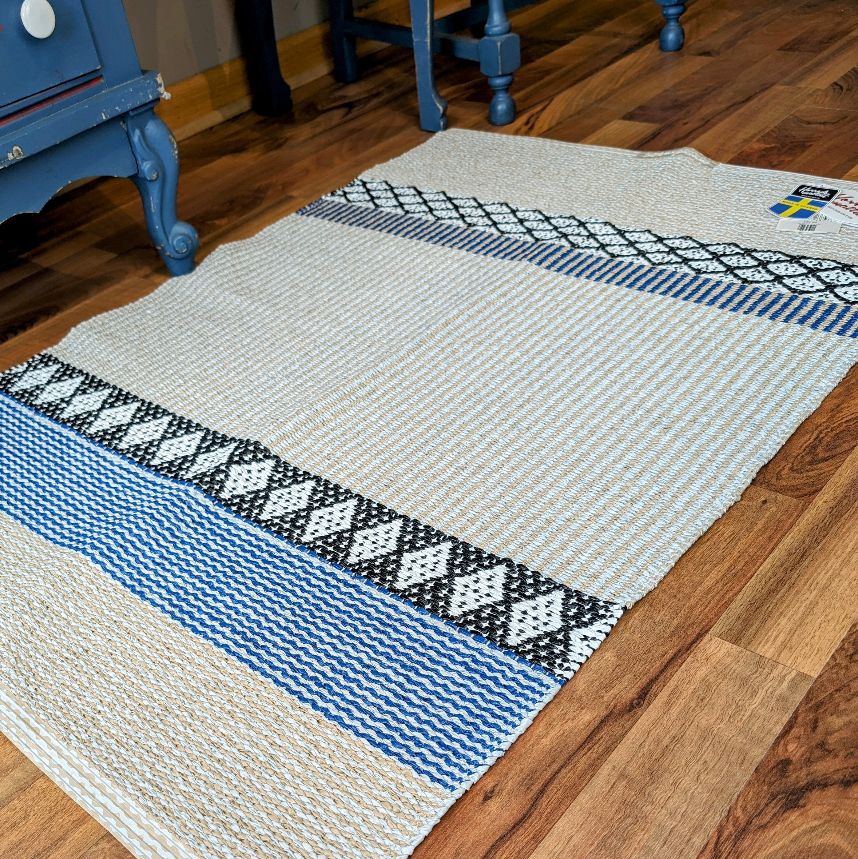 A Rug: Savanne Blue Vinyl with blue, white, and black geometric patterns rests on a wood floor, partially tucked under a blue chair. This stylish piece is maintenance-free, offering both elegance and ease in your living space.