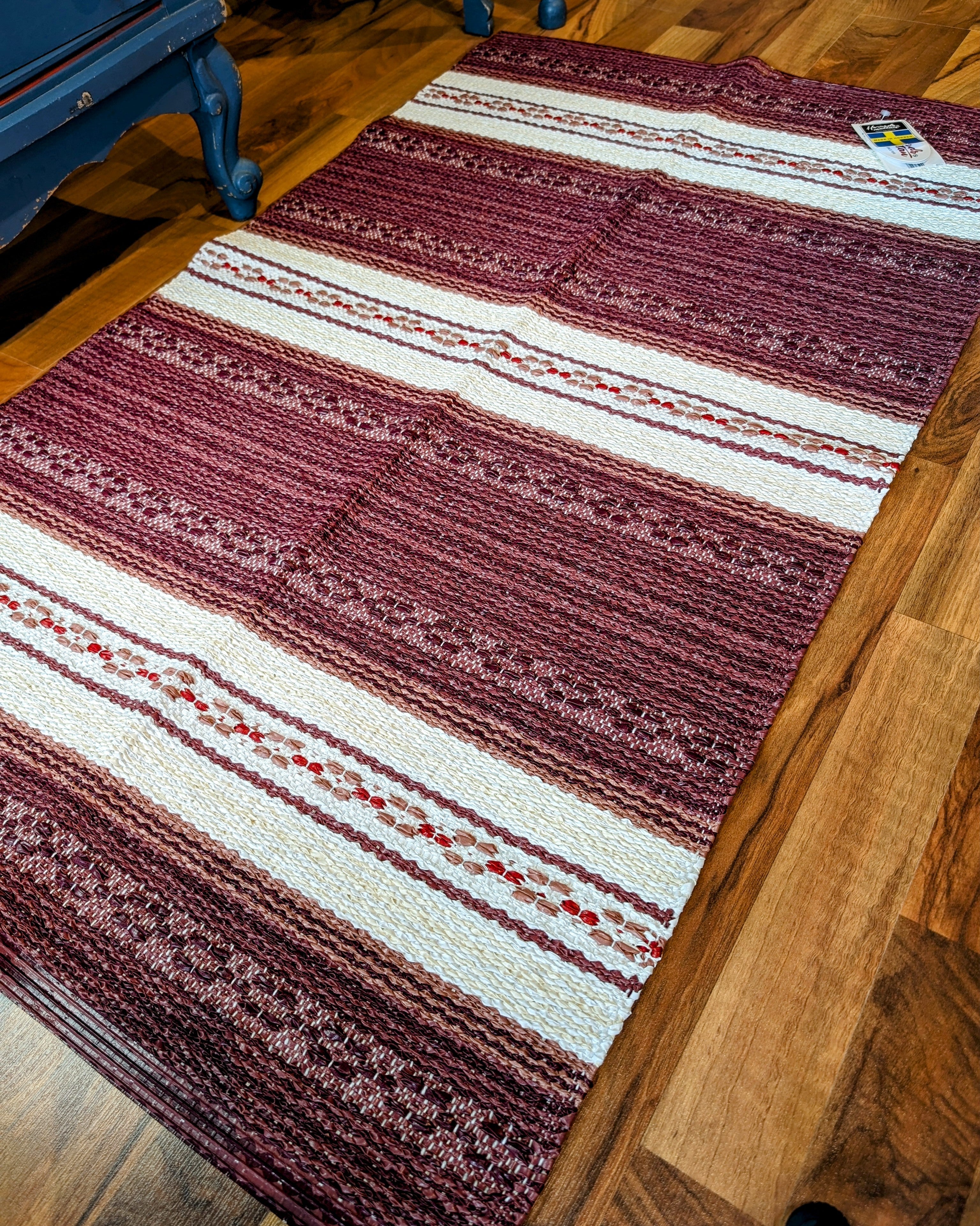 The Vinyl Rug: Horredsmattan - Astor - Red, featuring decorative maroon and white stripes, is laid out on a wooden floor.