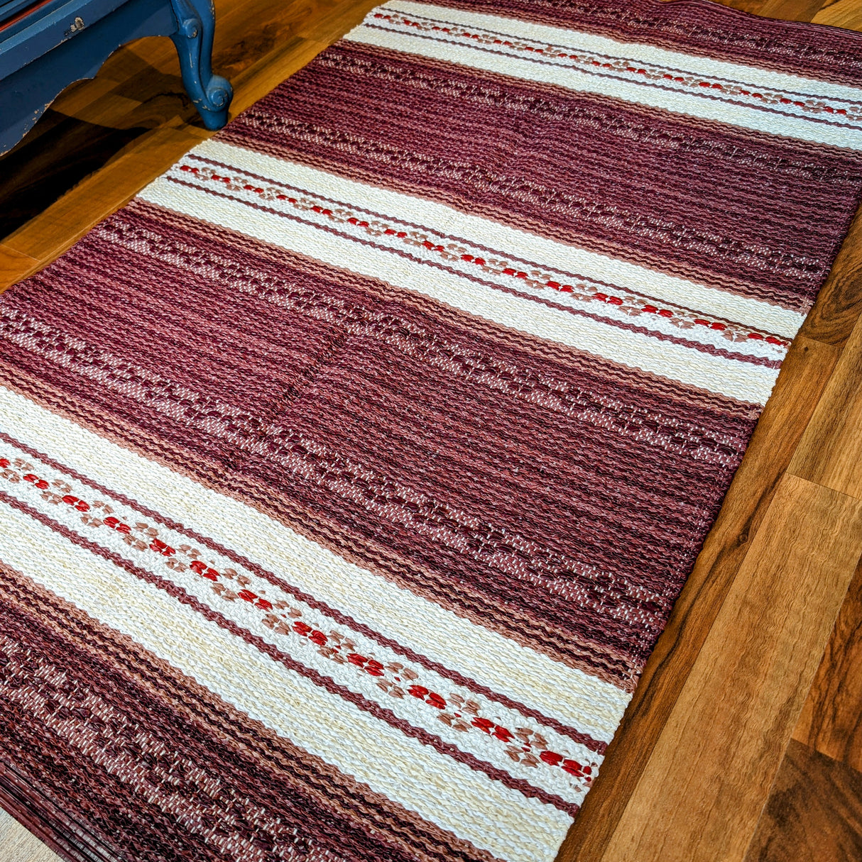 A Swedish vinyl rug, the Horredsmattan Astor in red, featuring maroon, white, and red stripes, lies on a wooden floor.