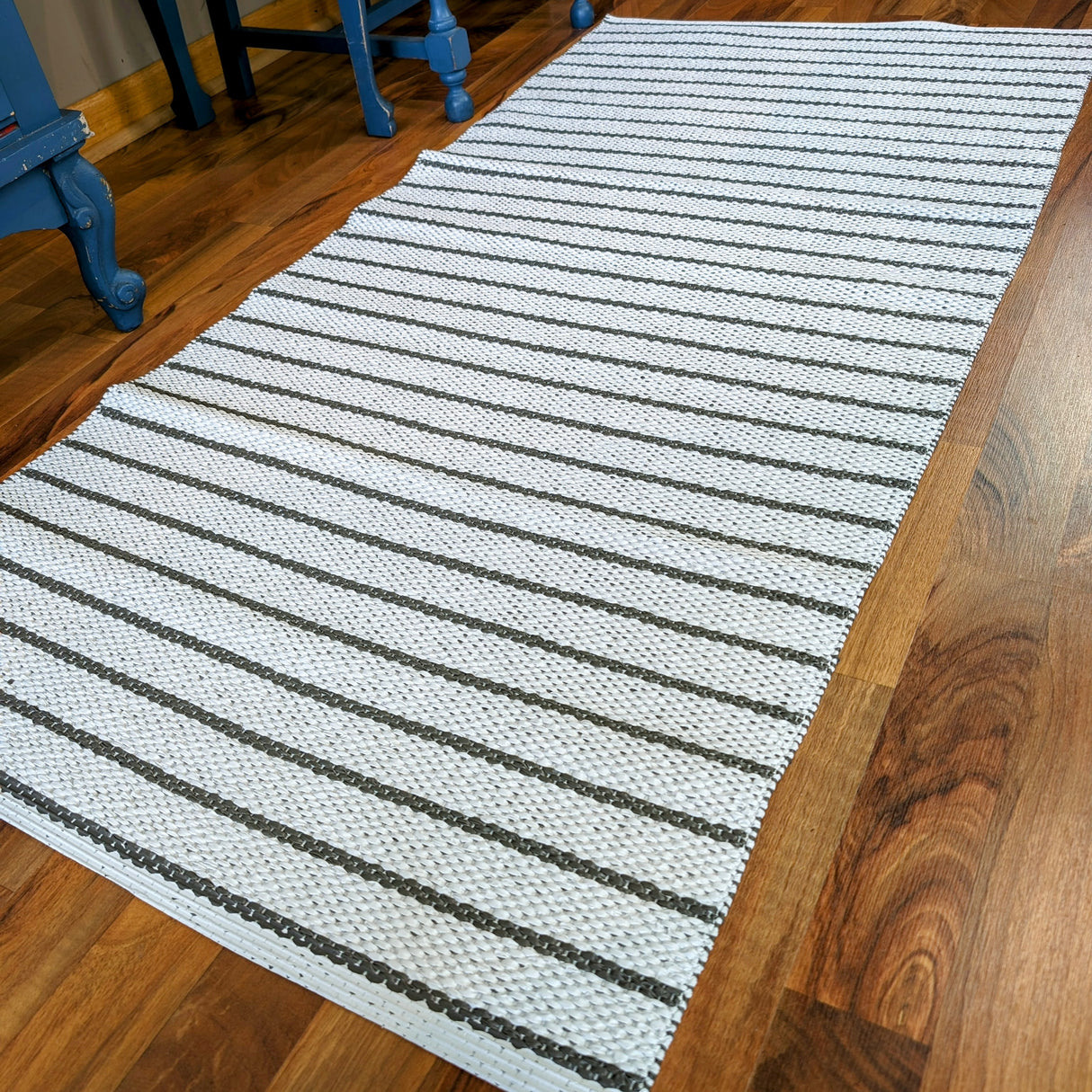 A dark gray woven vinyl rug, Horredsmattan - Music, lies on a wooden floor near blue furniture legs.