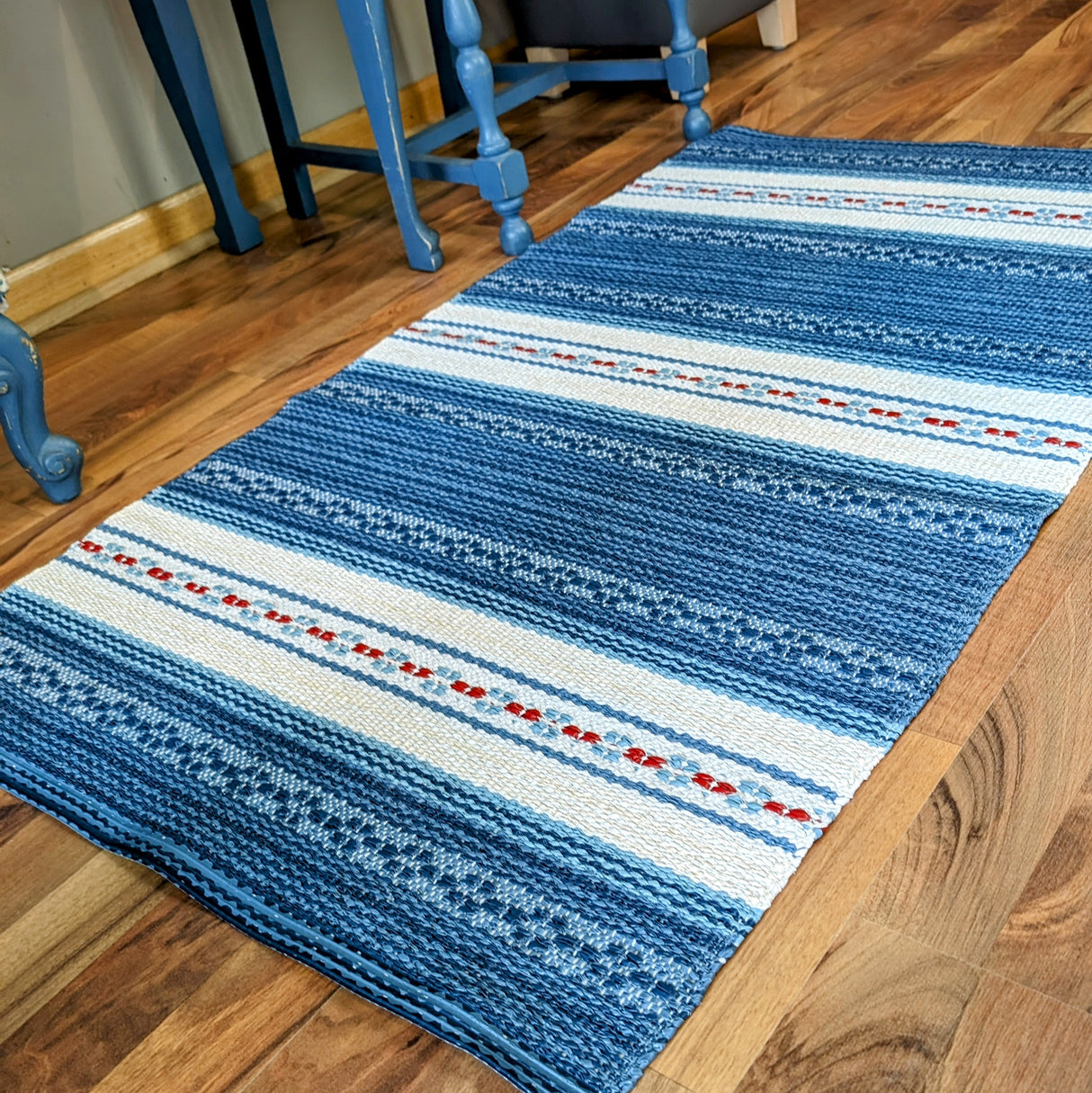 An Astor Blue rug, featuring a woven blue and white pattern with red accents, lies on a wooden floor next to blue furniture. This maintenance-free, durable piece is renowned for its Swedish vinyl design and adds charm and resilience to any space.