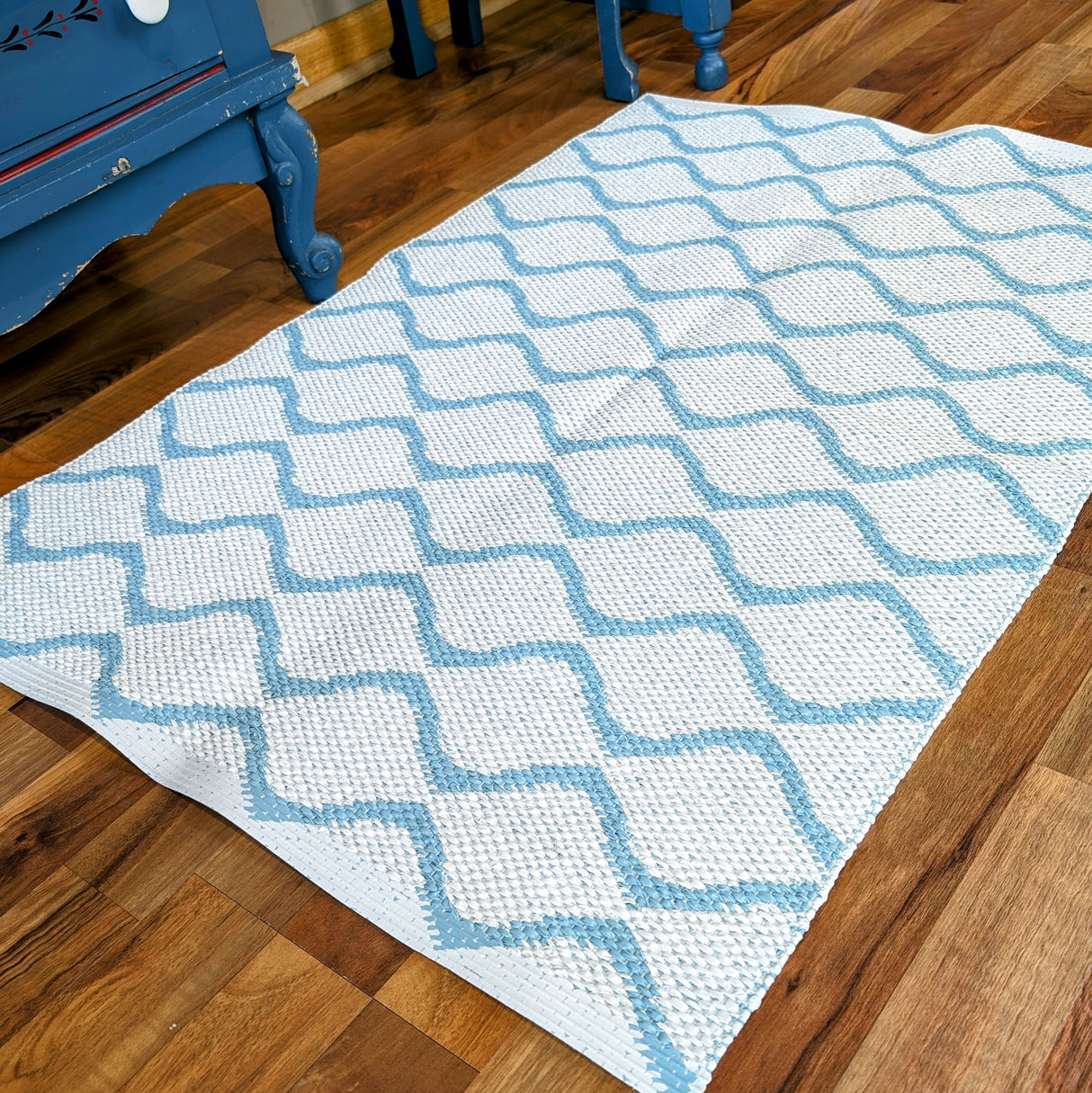 The Wave Blue rug, featuring a durable and long-lasting white weave with a blue lattice pattern, lies on a wooden floor next to a blue piece of furniture.
