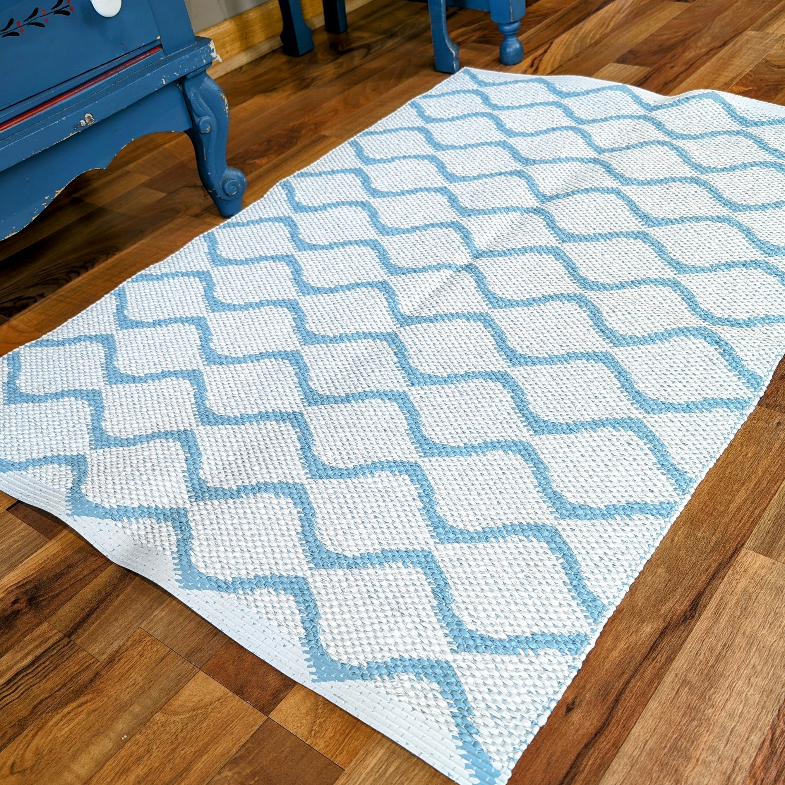 The Wave Blue rug, featuring a durable and long-lasting white weave with a blue lattice pattern, lies on a wooden floor next to a blue piece of furniture.