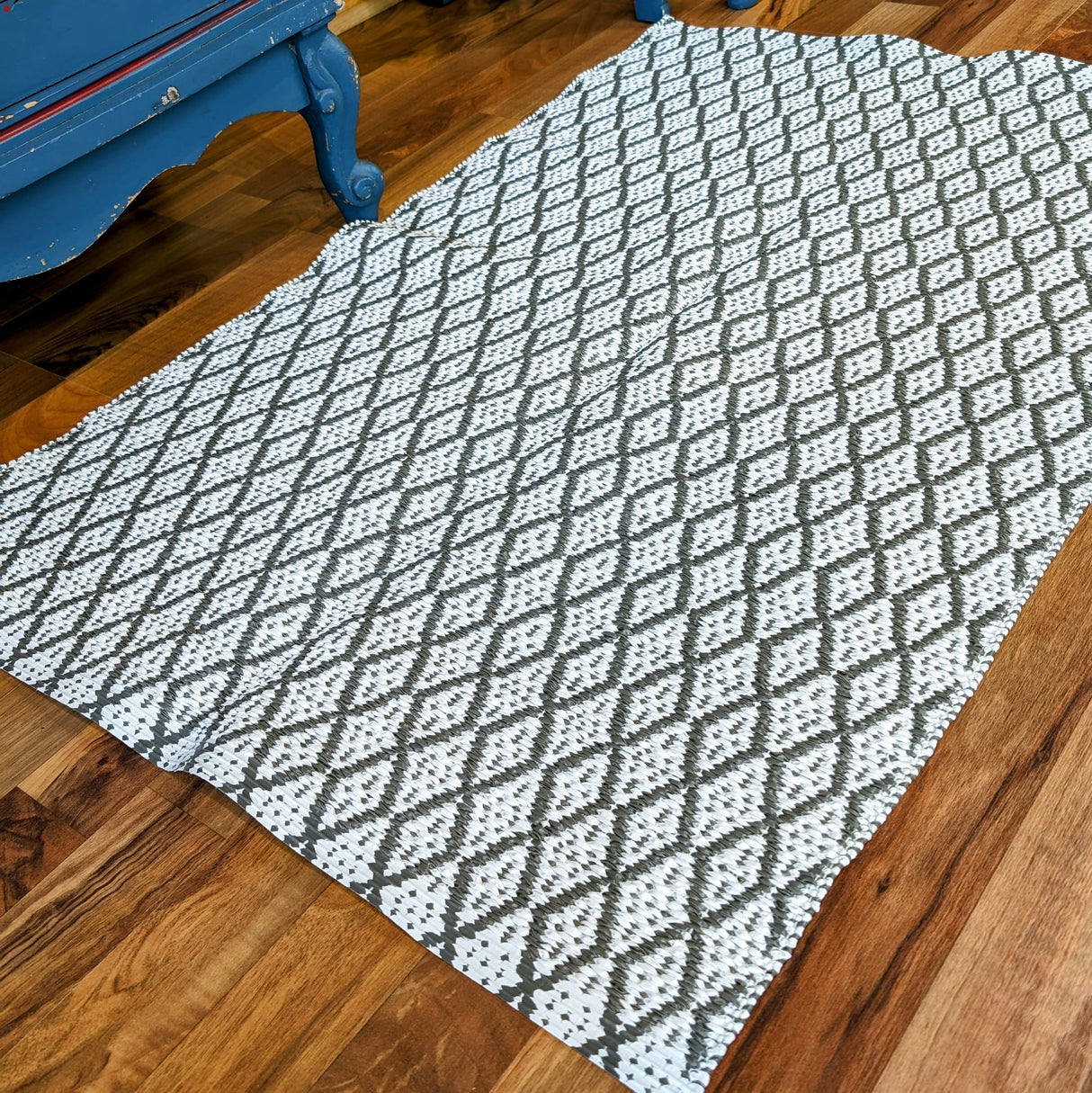 The Eye Grey 70x100cm rug, featuring a durable, maintenance-free woven design with a black and white diamond pattern, complements the wooden floor next to a piece of blue-painted furniture.