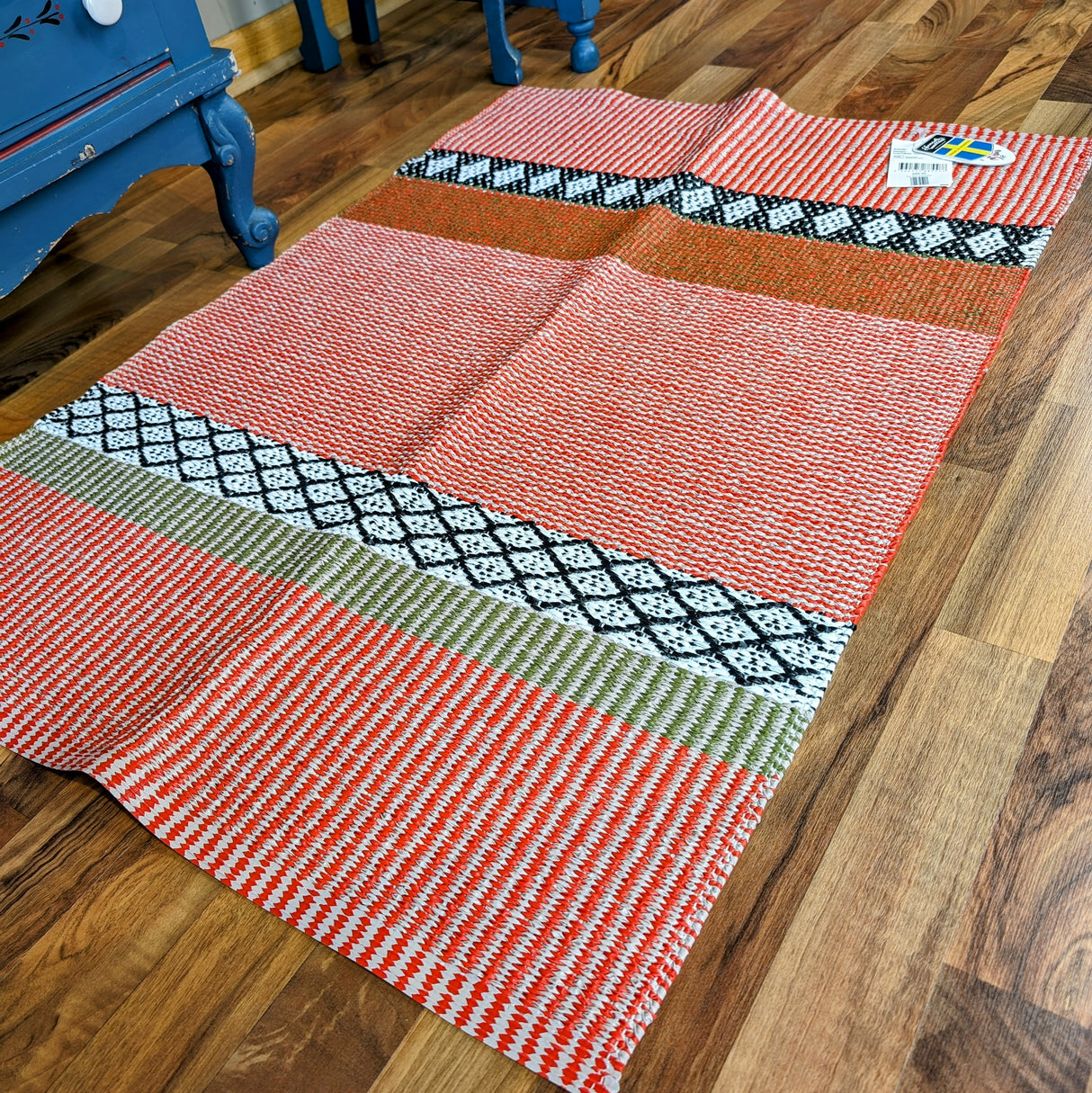 The Horredsmattan Savanne vinyl rug in reddish orange, beige, white, black, and green geometric patterns lies on a wooden floor by a blue piece of furniture. A price tag is attached to one corner of this machine washable rug.