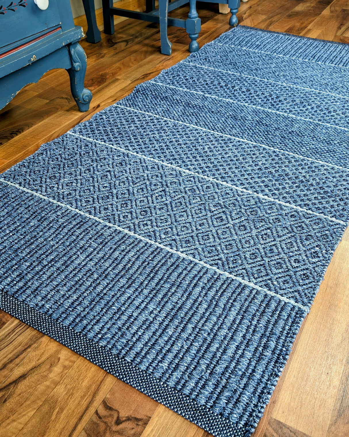 A Mixed Alice rug in blue, made of cotton and vinyl, featuring a textured Swedish design with a geometric pattern, lies on a wooden floor near blue furniture, making it ideal for both indoor and outdoor settings.