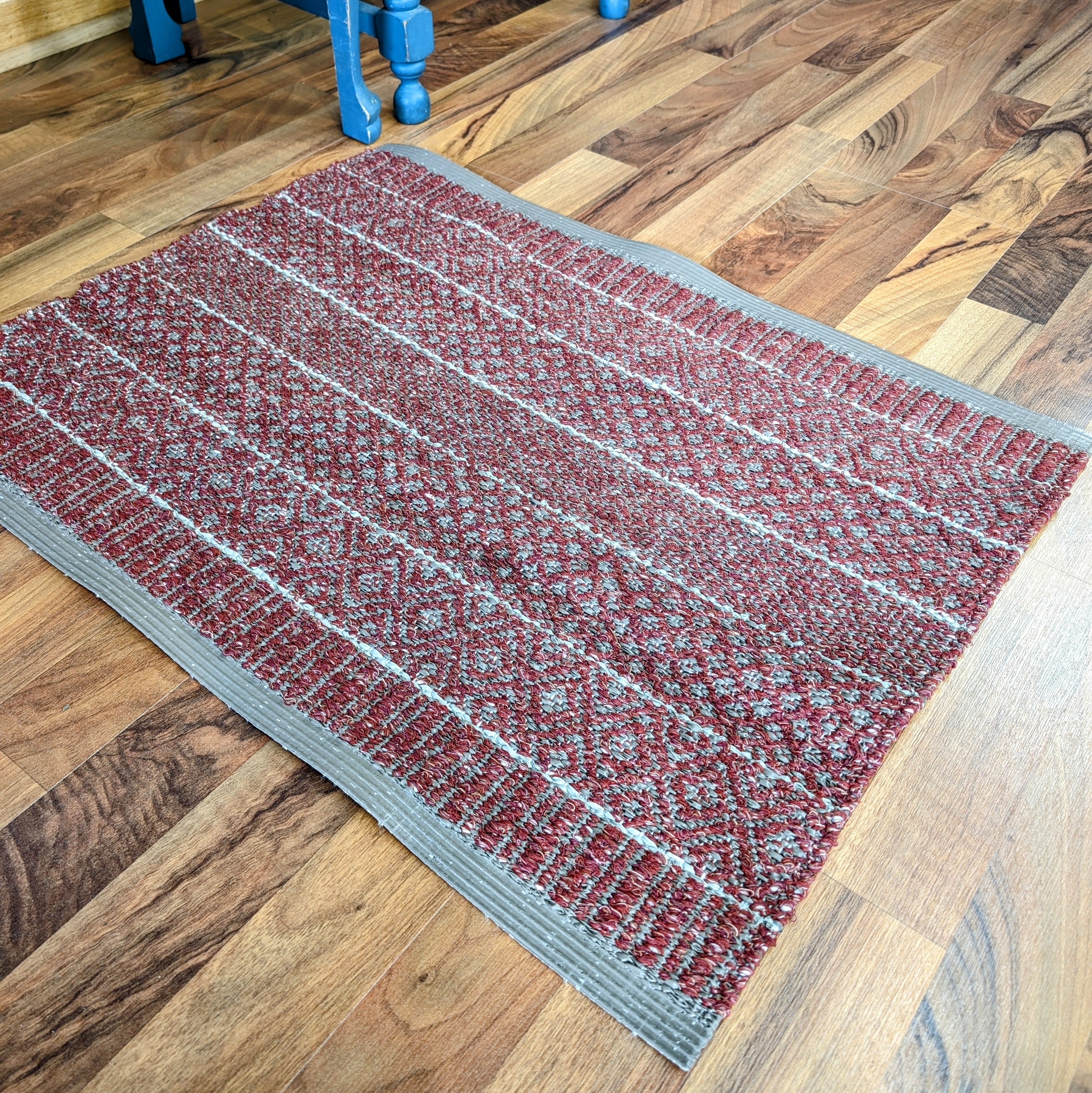 A Mixed Alice burgundy rug, featuring a cotton and vinyl blend with a geometric pattern, sits elegantly on the wooden floor, perfect for indoor and outdoor use.