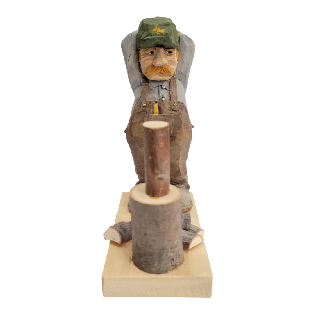 The Woodchopper figurine by Bill Erickson features a hand-carved man in a green hat and overalls, standing behind a chopping block with logs, hands relaxed behind his head, showcasing rustic charm.