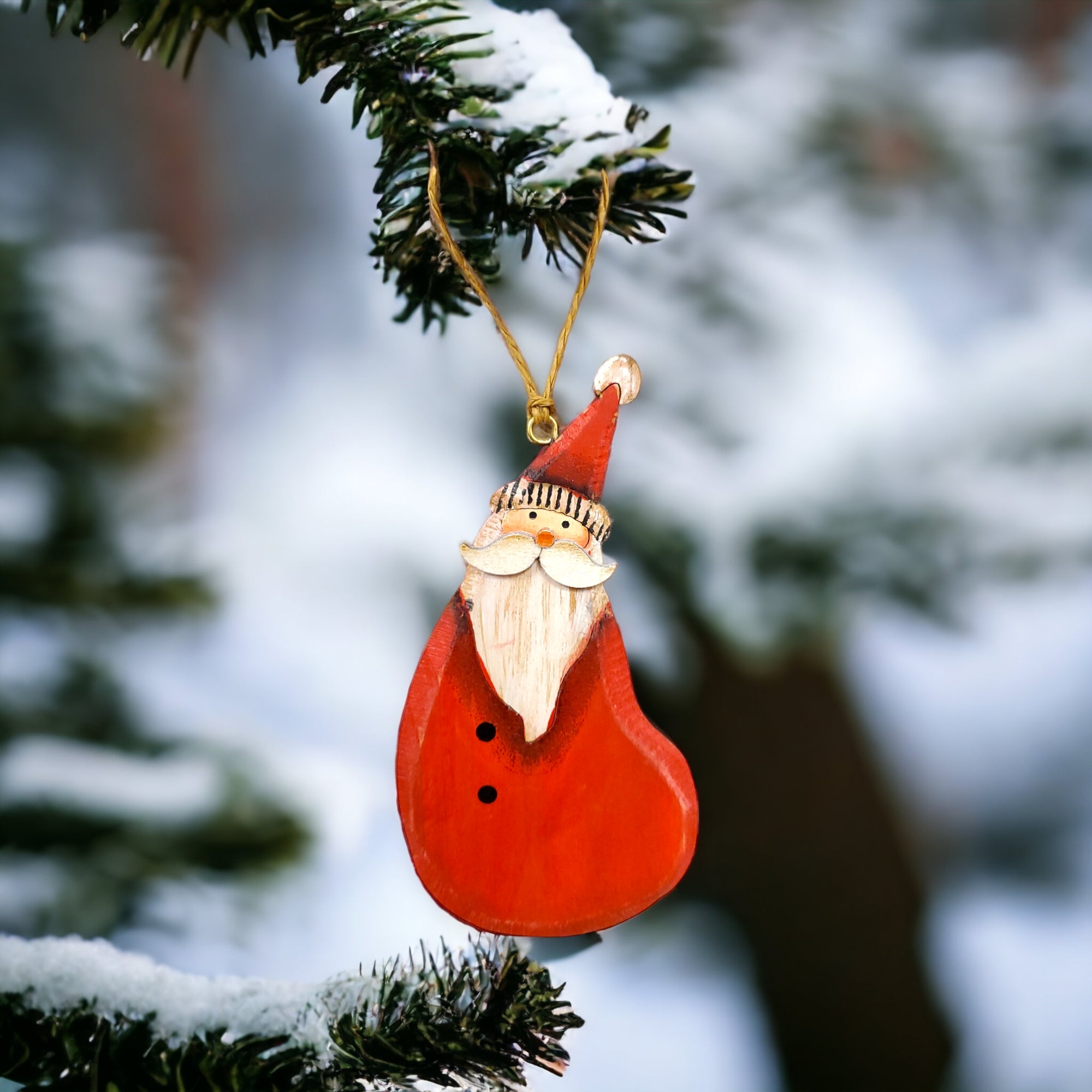Close-up of a Christmas tree ornament called "Ornament: Country Santa," featuring a Wooden Santa with a 3-dimensional nose, hanging on a pine tree branch against a snowy background.