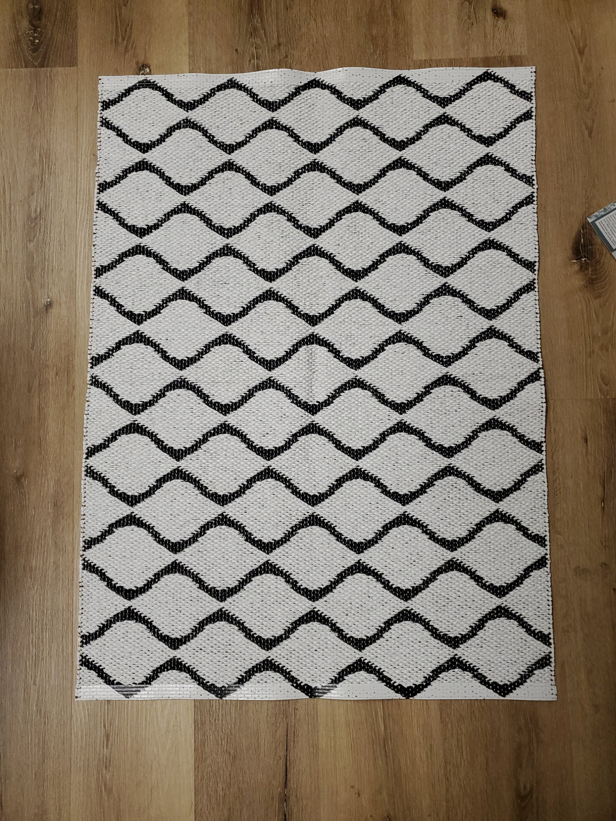 The Rug: Wave Black, a rectangular Swedish vinyl rug featuring a black wavy line pattern, adds durable and long-lasting appeal to a wooden floor.