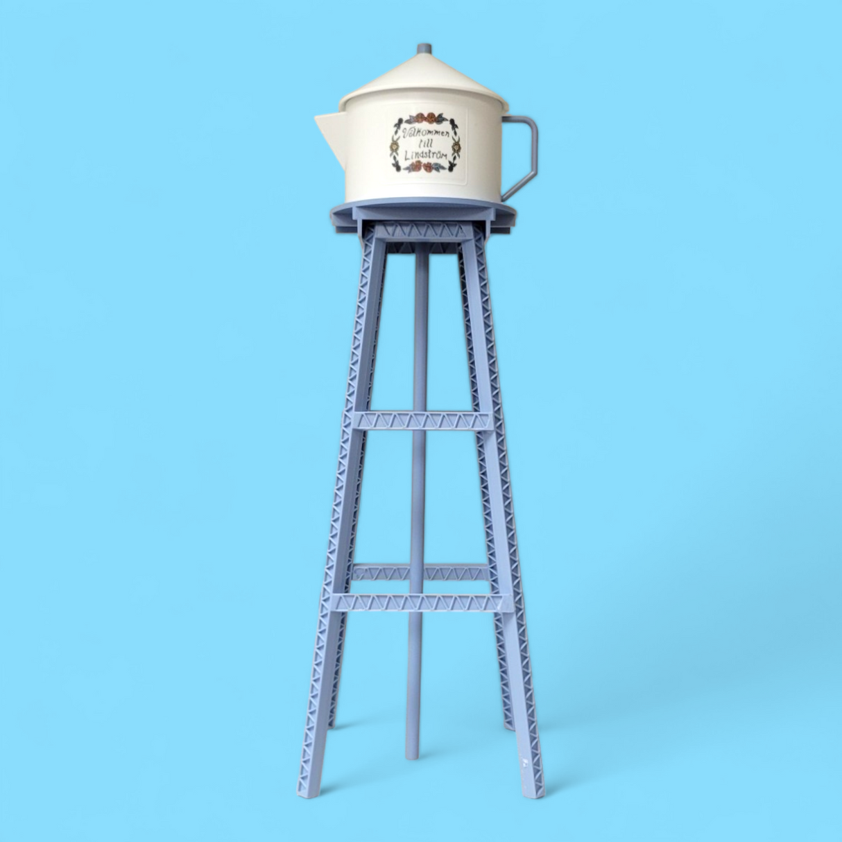 A Model Kit: Miniature Replica of the Lindström Coffee Pot Water Tower features floral designs and sits atop a tall blue metal structure, set against a solid light blue background.