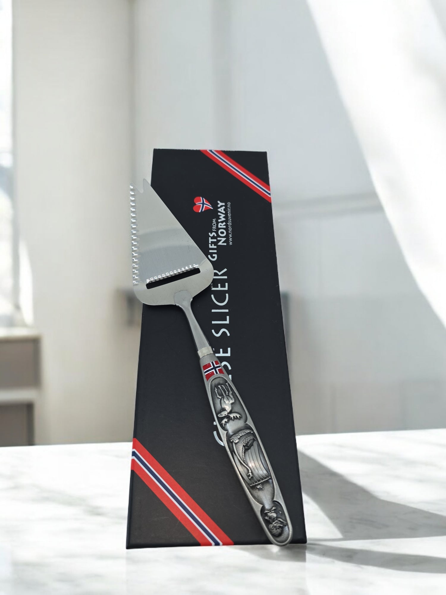 A Norwegian Cheese Slicer featuring images of a Viking ship and moose on its pewter handle stands before a black GIFTS FROM NORWAY box decorated with red, white, and blue stripes.