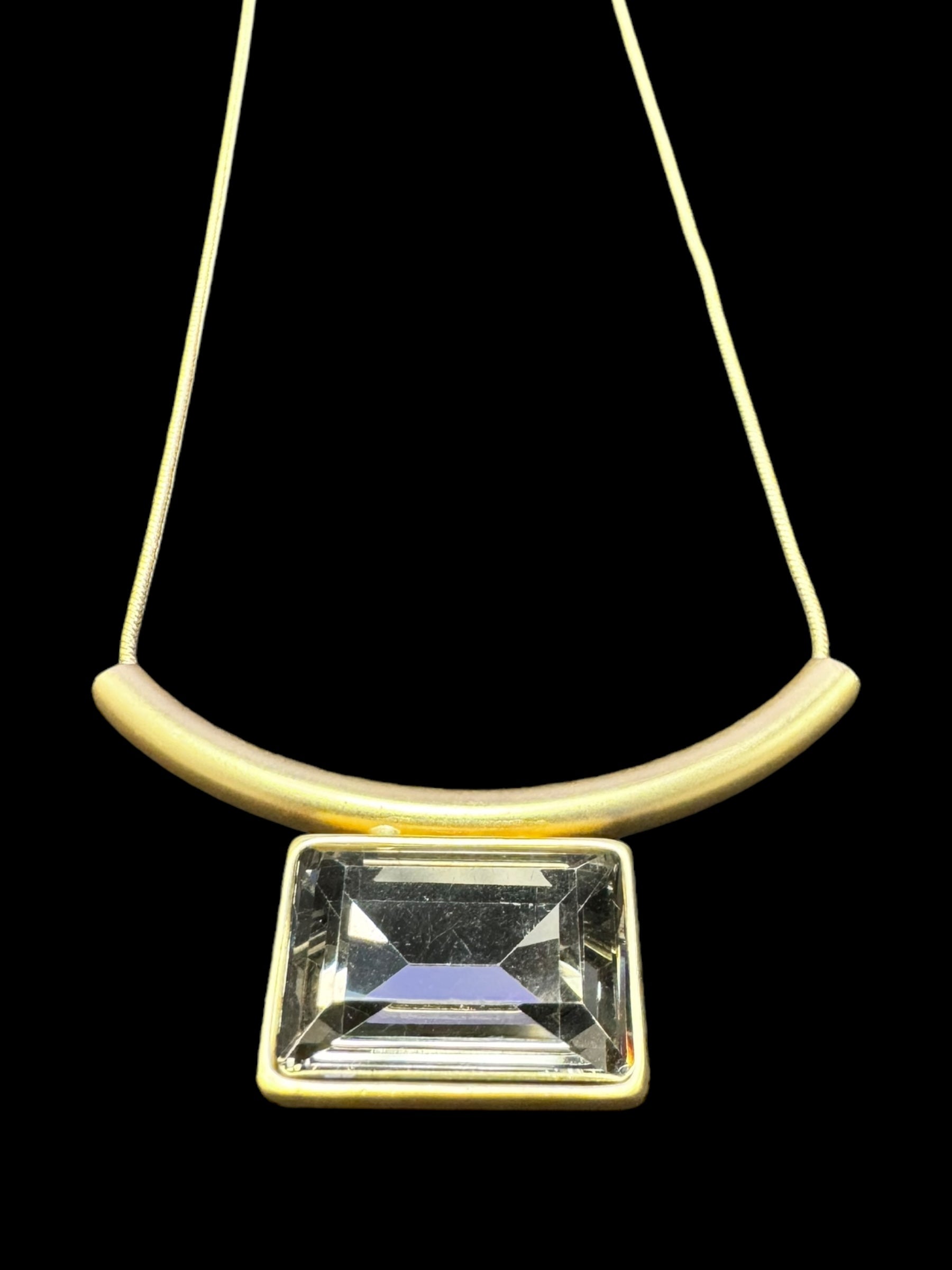 The Necklace: Audrey Black Diamond Gold Plated by DANSK Copenhagen exudes timeless elegance with its minimalist design featuring a curved gold bar, large rectangular gem, and thin chain set against a black backdrop. Its simplicity makes it a versatile accessory for any occasion.