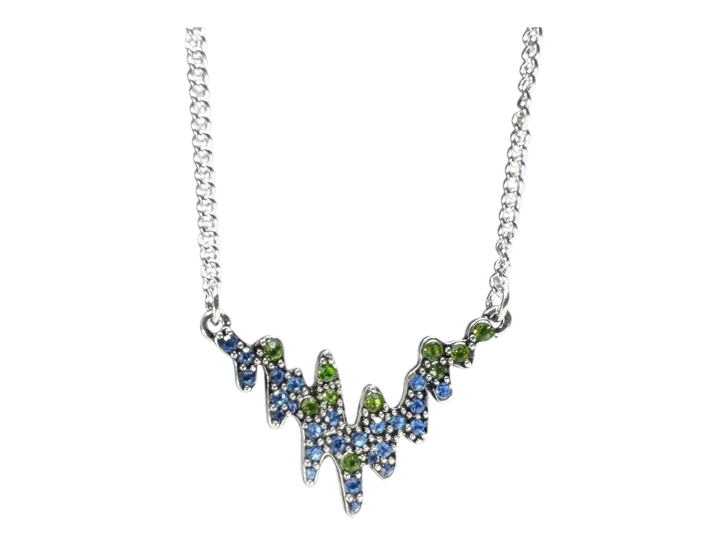 The Necklace: Northern Lights A&C Oslo Norway features a silver plated chain with a jagged V-shaped pendant, adorned with blue and green crystals, inspired by Norwegian jewelry and reminiscent of the aurora borealis.