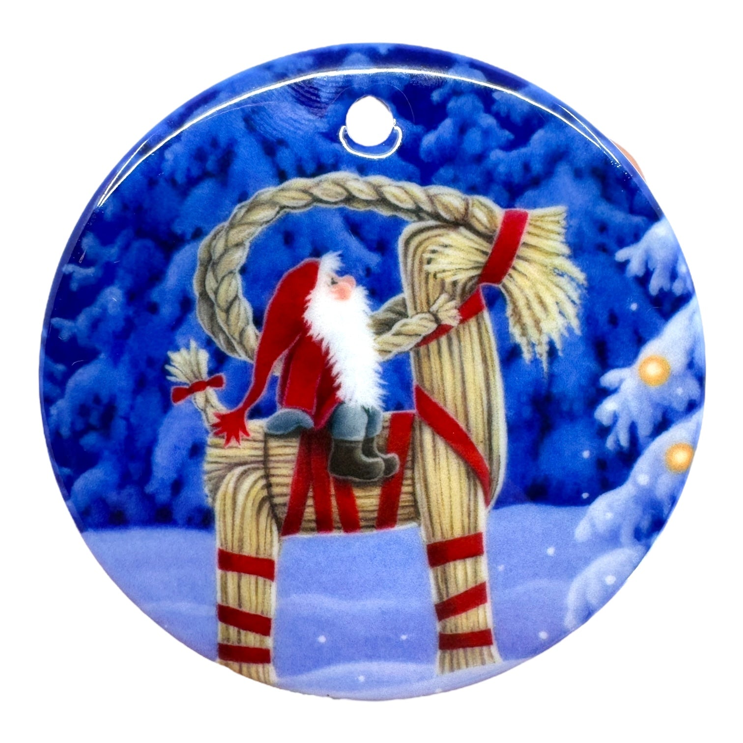 The 3" round ornament "Tomte on Straw Goat" by Eva Melhuish features a Scandinavian Tomte in a red hat sitting on a straw goat, set against a snowy backdrop with a decorated tree.