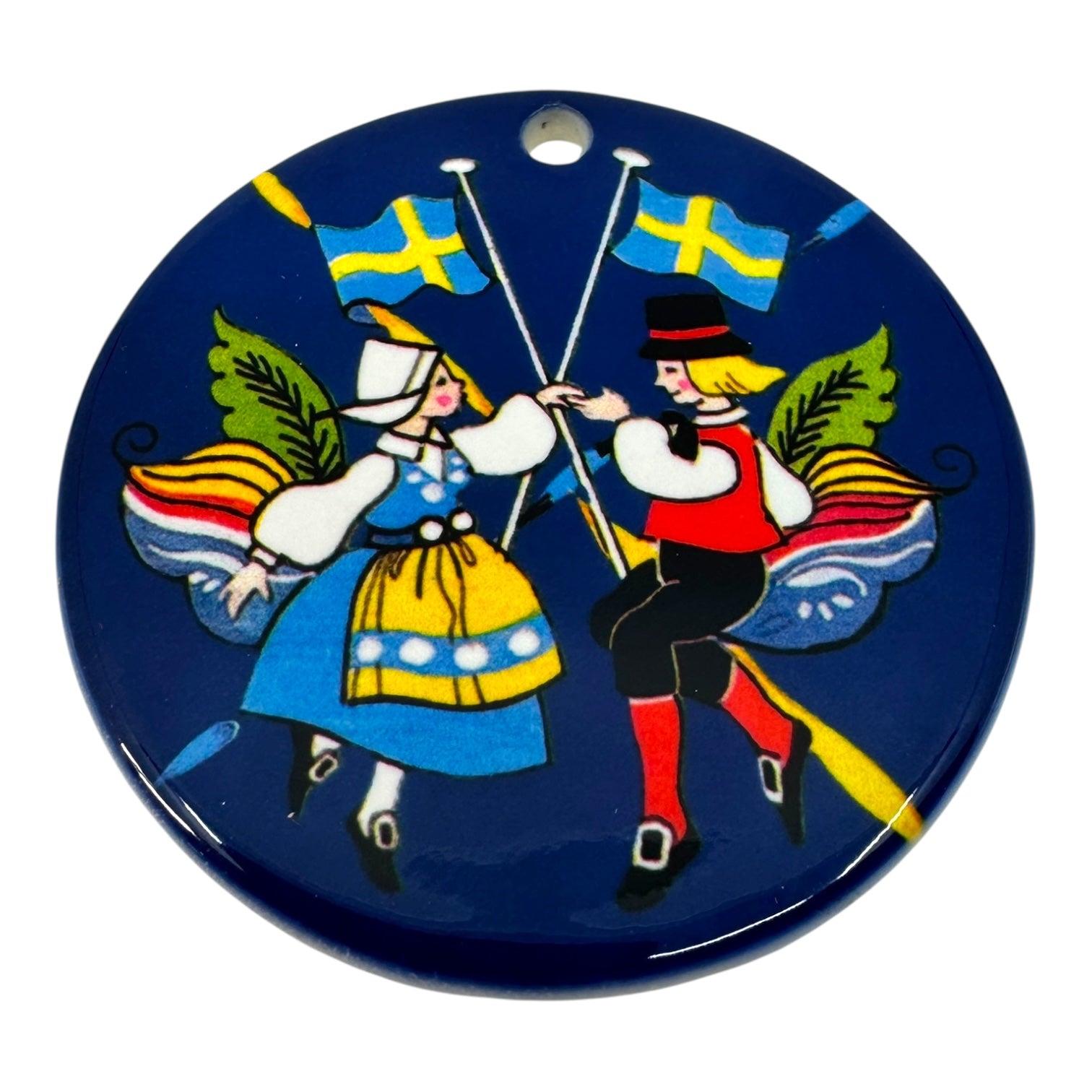 Ornament: Swedish Dancers – a round blue porcelain ornament displaying dancers in Swedish folk costumes, with crossed flags and vibrant wings in the background, capturing the charm of Scandinavian culture.