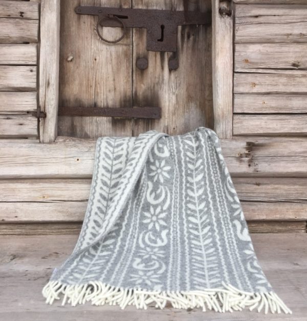 The luxurious Anno 1862 blanket by Kerstin Landstrom, crafted from 75% Norwegian lambs wool, 15% Merino wool, and 10% silk in gray and white with fringes, is elegantly draped over a wooden step in front of a rustic door.