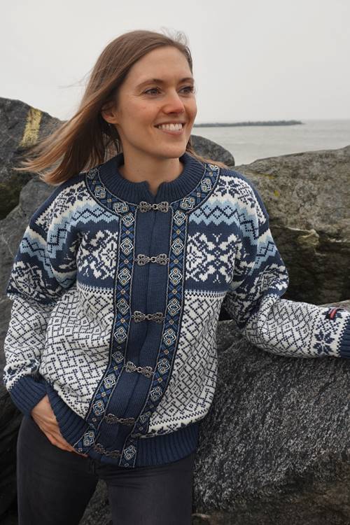 A person wearing the Gjestal - Classic Norwegian Blue Cardigan with genuine pewter buckles smiles while standing against a rocky backdrop near the sea.
